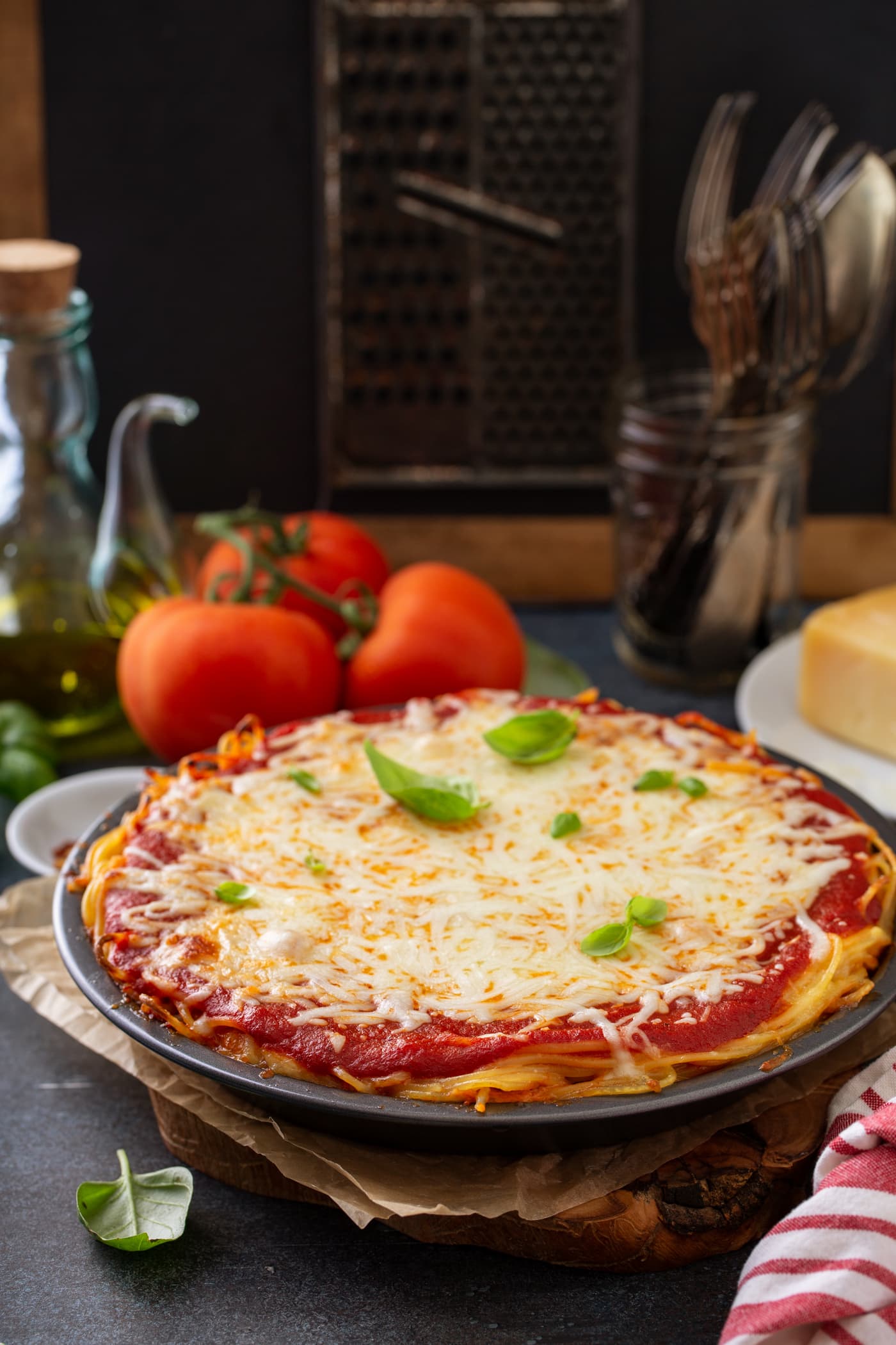 A pie dish filled with baked spaghetti pie. There are layers of spaghetti noodles, spaghetti sauce and melted cheeses. There are a few basil leaves on top and three tomatoes, olive oil, cheese, and a jar of forks in the background.