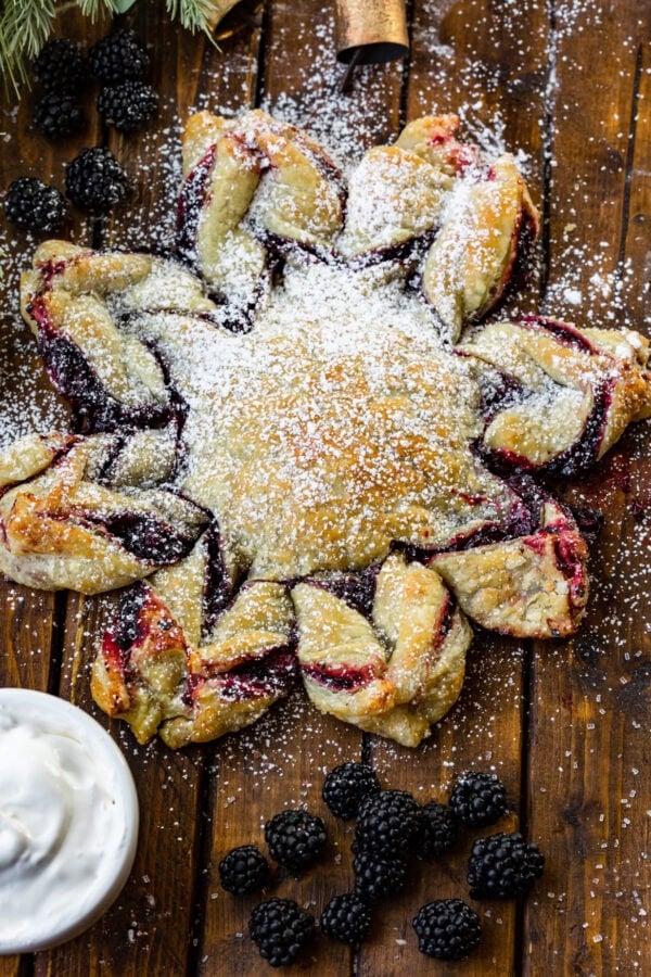 a photo of a blackberry puff pastry snowflake dusted with powdered sugar with whole blackberries scattered around it