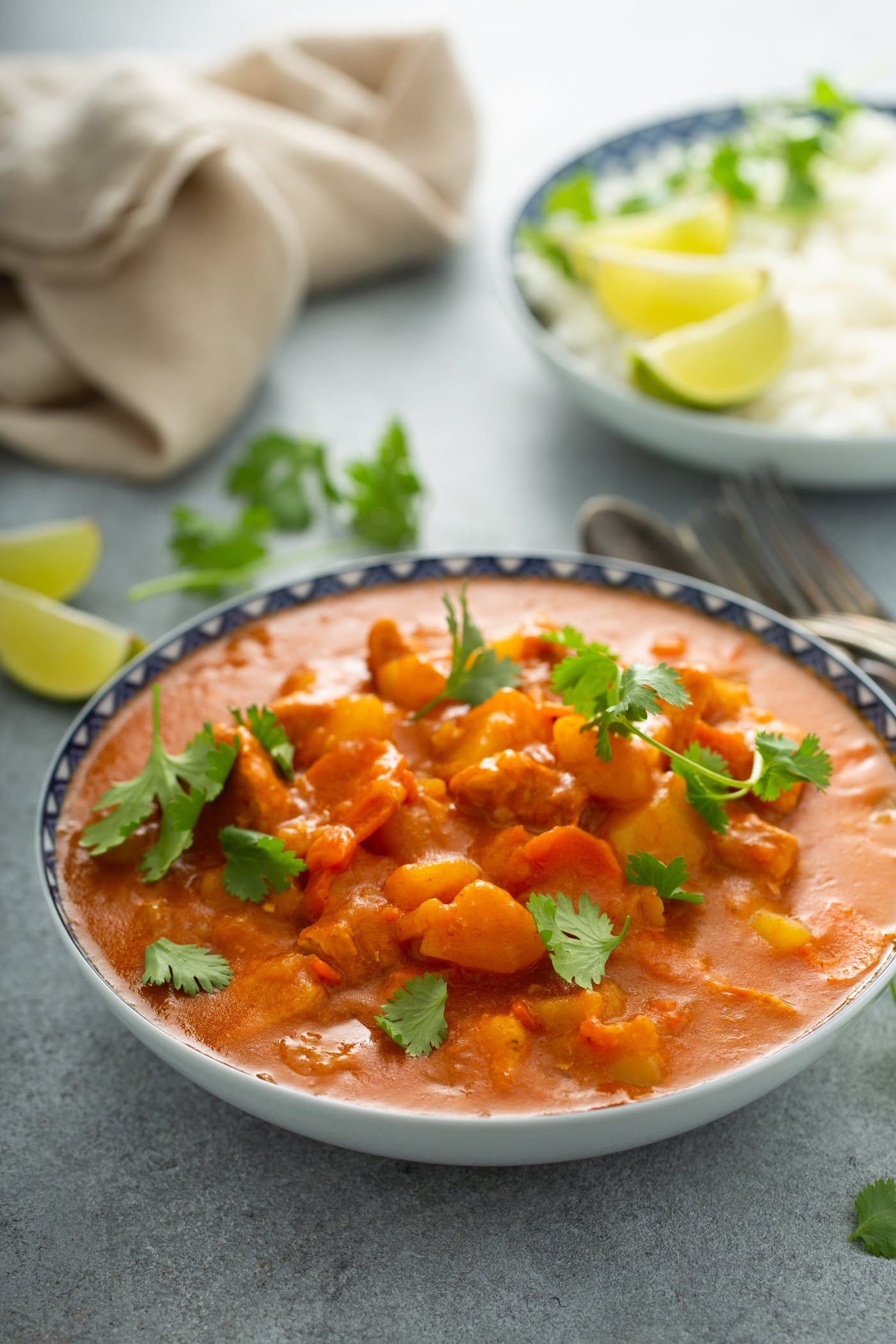 A large bowl of chicken curry. There are chunks of chicken and potatoes in a bright curry sauce. Parsley leaves and lime wedges are in the background, and a fork and spoon are next to the bowl of curry.