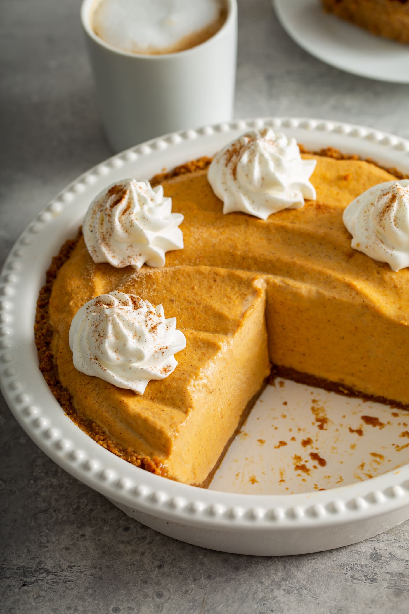 A white pie dish with a pumpkin pie ice cream pie in it. A slice has been removed from the pie and the remainder of the pie is topped with dollops of whipped cream sprinkled with cinnamon. A mug of hot cocoa topped with whipping cream is in the background.