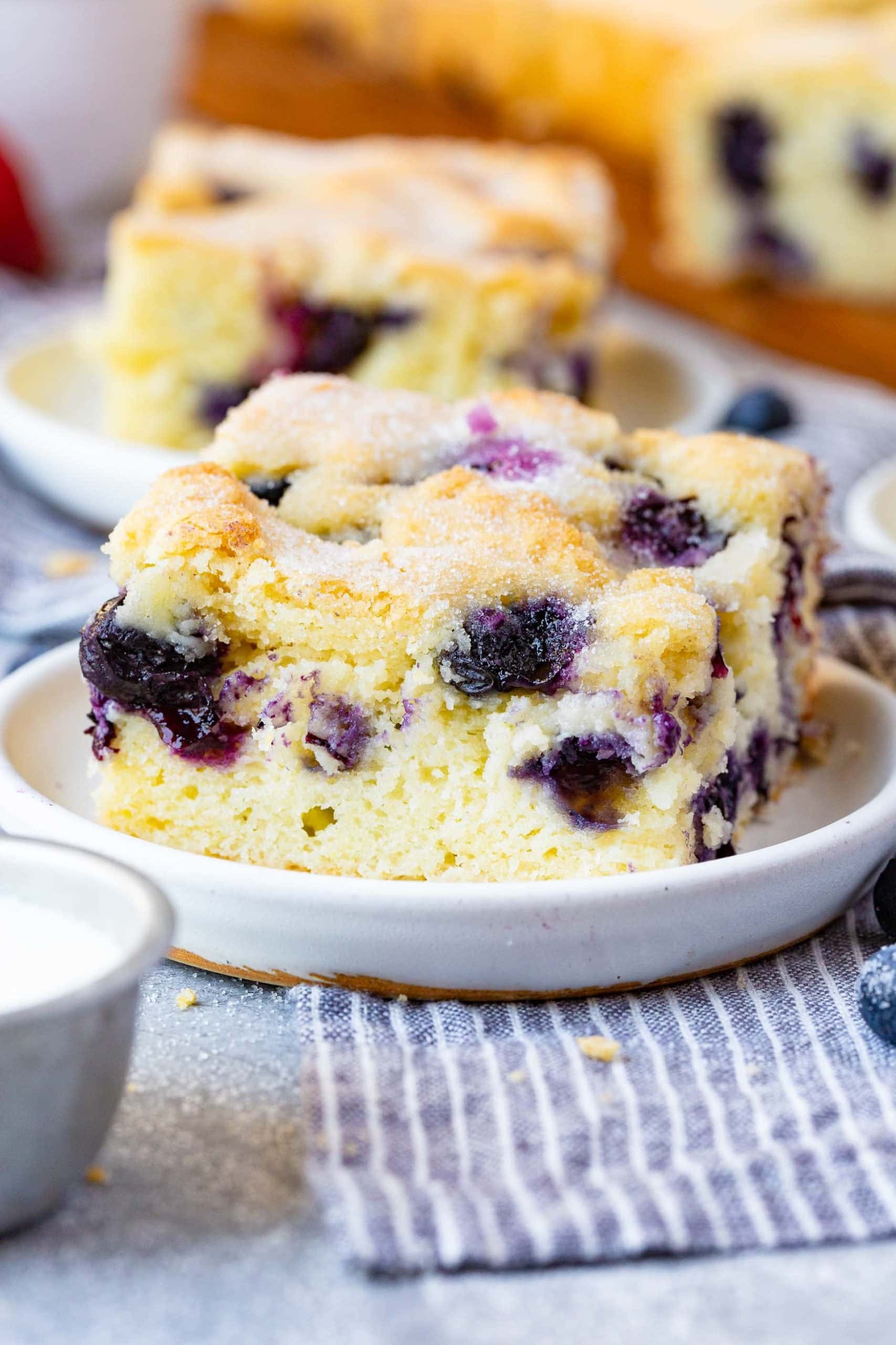 a photo of a piece of blueberry breakfast cake on a small white plate full of purple fresh blueberries.