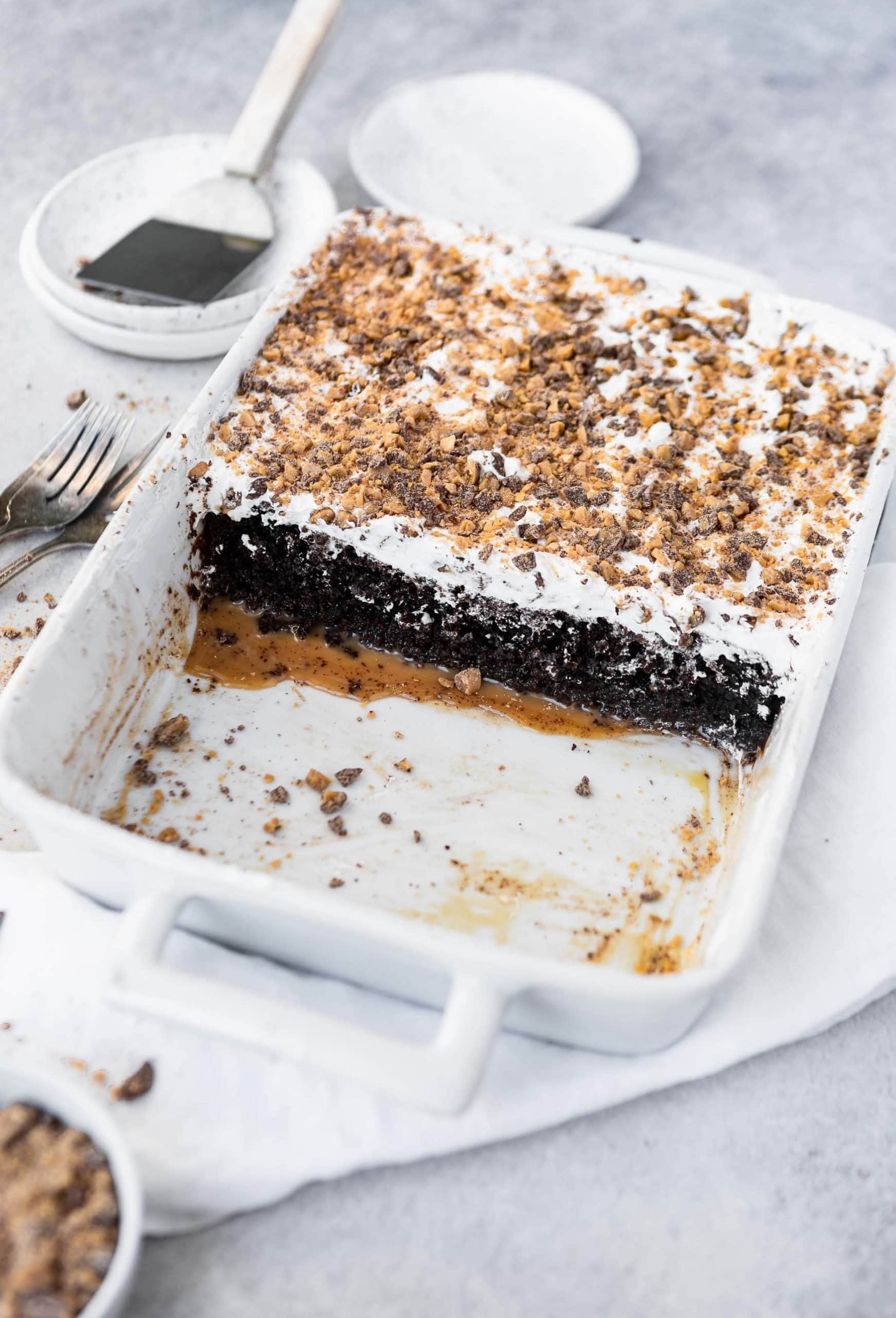a photo of a white baking dish half full of a chocolate cake topped with whipped cream and toffee bits. the cake is oozing caramel out the bottom into the empty half of the pan.