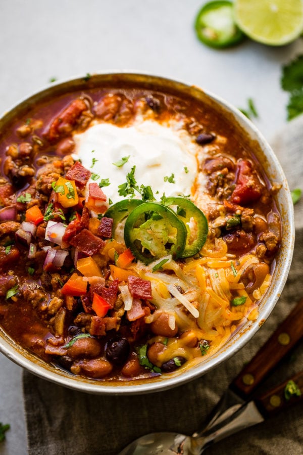 Award winning healthy turkey instant pot chili A metal bowl of ground turkey chili full of beans, red onions that have been chopped, chopped peppers , jalapeno slices on melted cheese next to a dollop of sour cream with a dash of cilantro and a grey napkin underneath