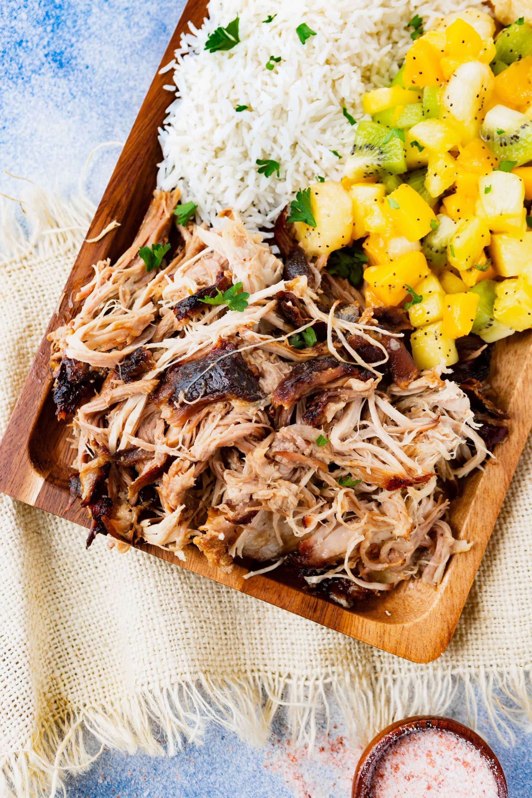 a photo of a rectangular wooden platter full of kalua pulled pork, herbed rice, tropical fruit salad and golden Hawaiian rolls.