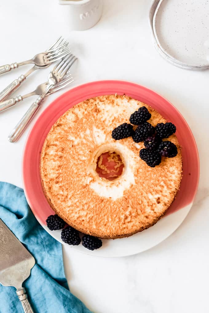 An angel food cake on a pink plate with blackberries on top and forks next to it.