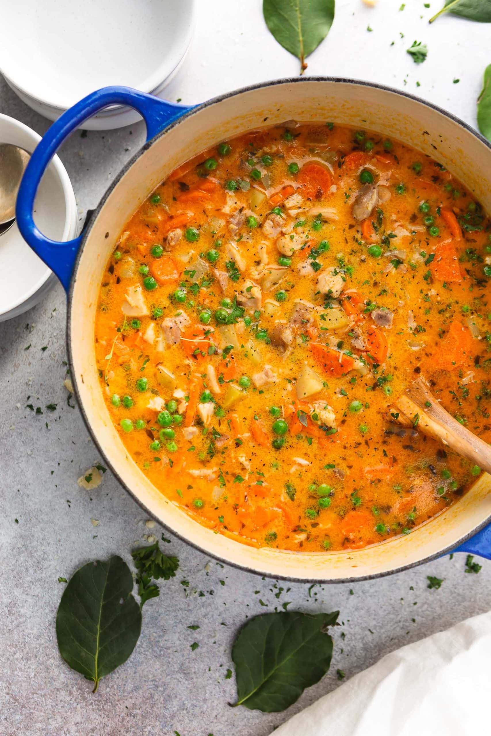 A large pot of slow cooker chicken stew. It is a bright yellow-red color and packed with bits of fresh herbs, vegetables and chicken. A wooden spoon is in the pot.