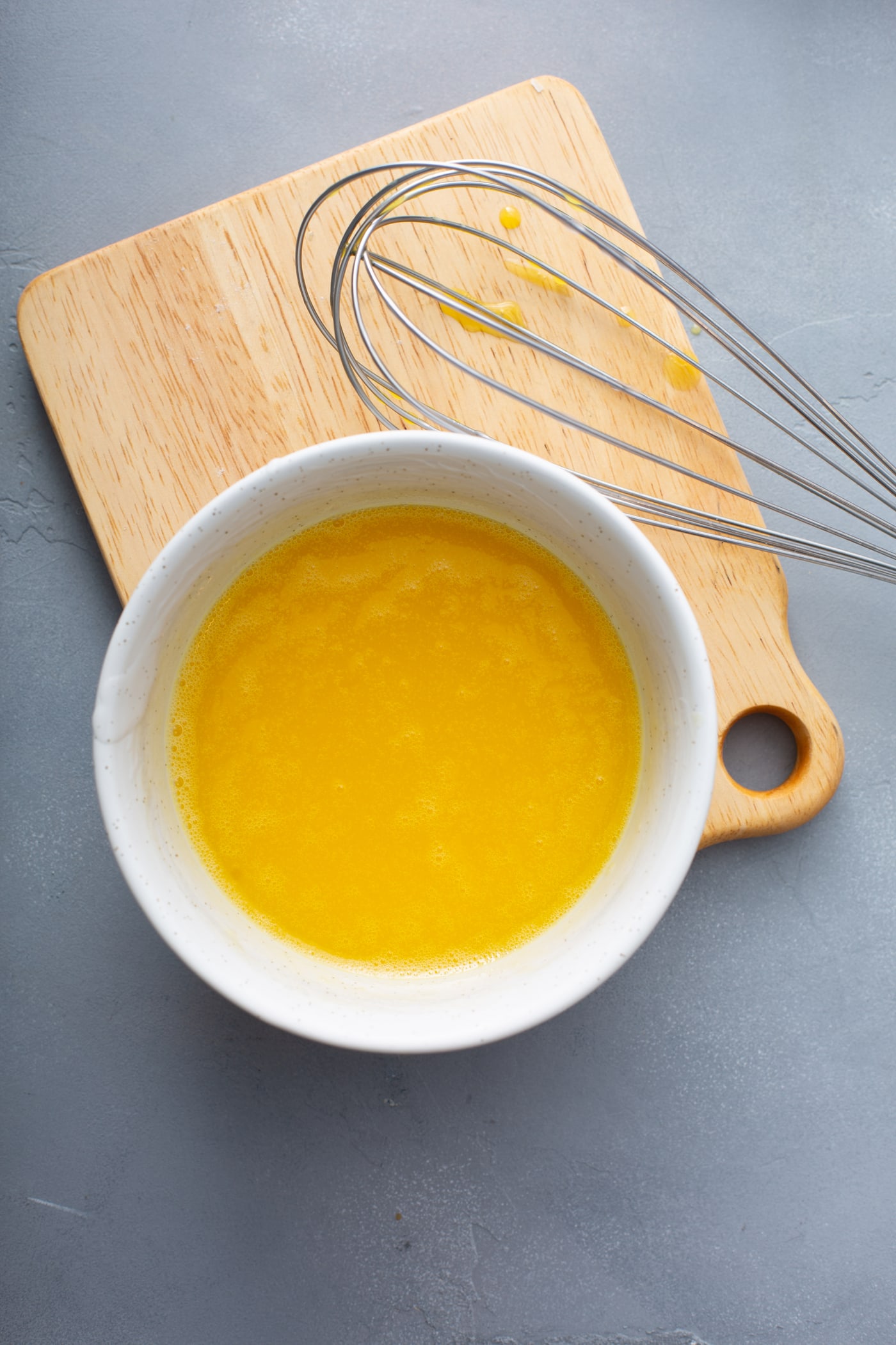 A bowl of egg yolks that have been whisked together. There is a wooden cutting board and a whisk by the bowl of egg yolks.