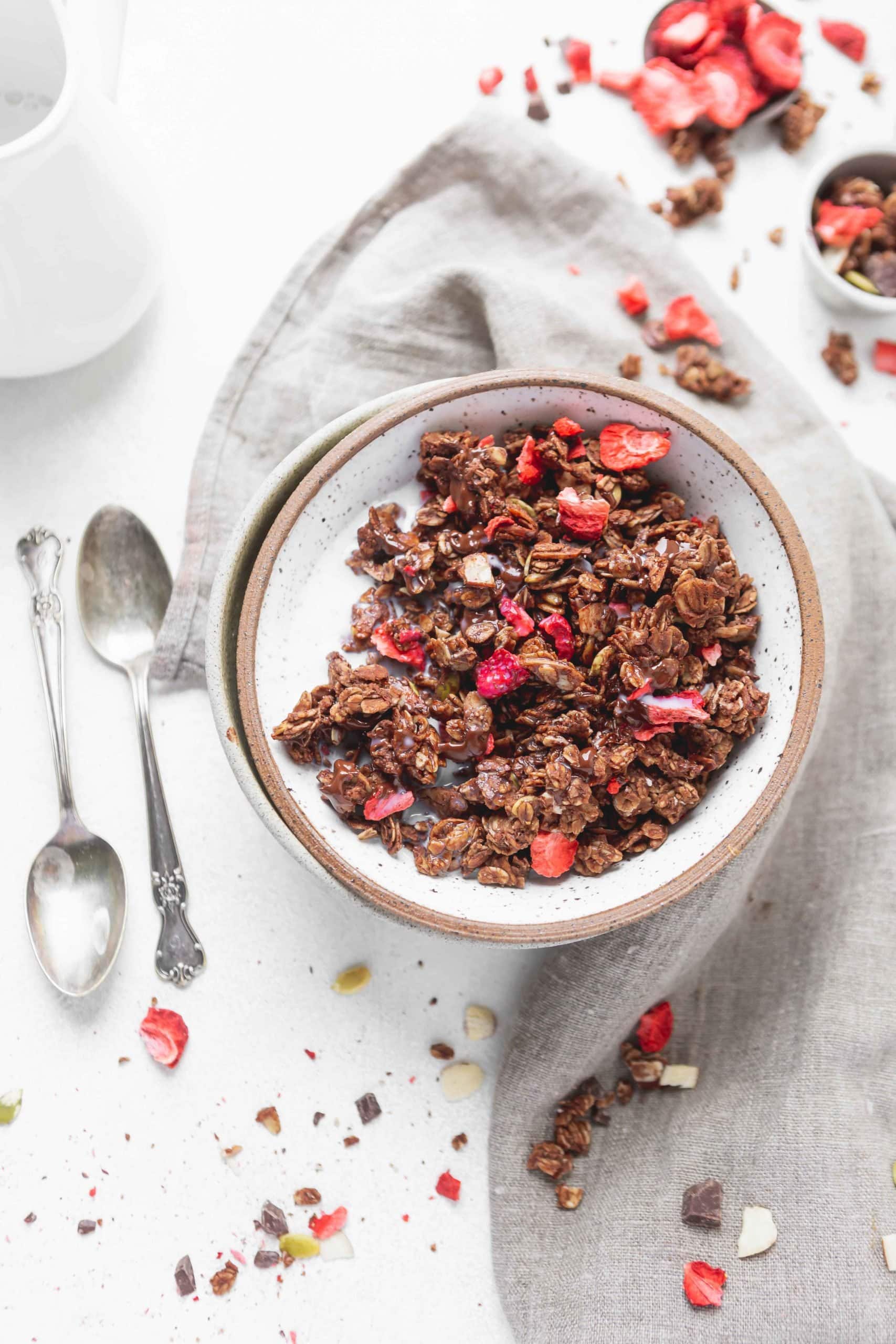 A bowl of chocolate lovers granola with strawberries. There are chunks of chocolate covered oats, chocolate pieces and strawberries. Two spoons are near the bowl.