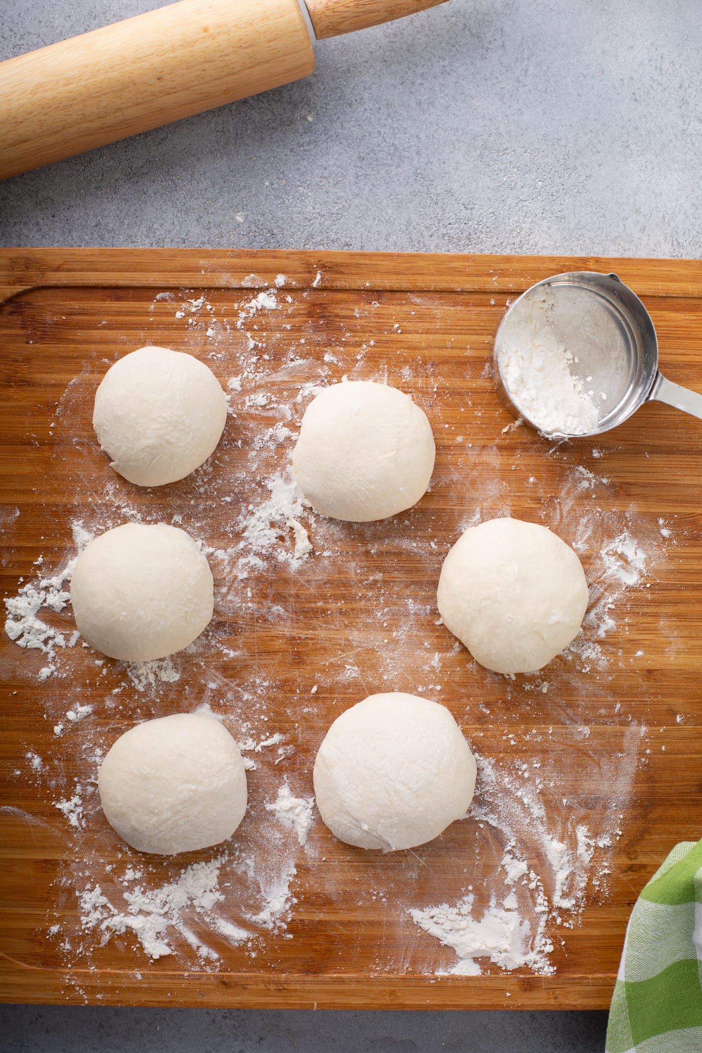 A photo of six blobs of unleavened bread dough sitting on a floured wooden cutting board with a measuring cup of flour sitting next to them.