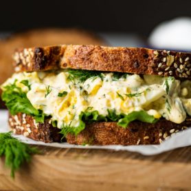 a photo of an egg salad sitting on a piece of parchment paper made on multigrain bread with lettuce and thick layer of egg salad.
