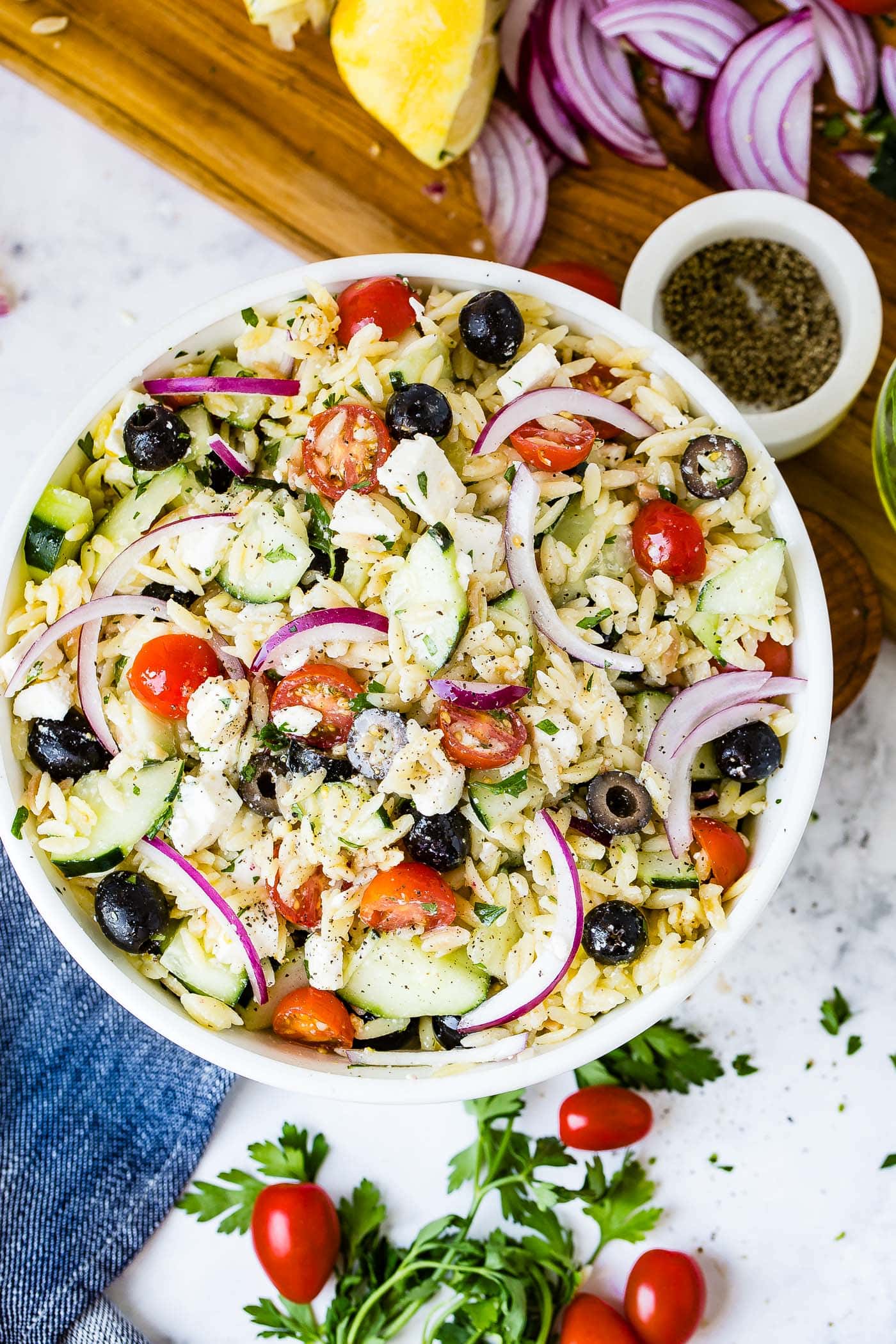 A white bowl of Greek orzo pasta salad with red onion, cherry tomatoes, feta cheese, cucumbers and black olives.