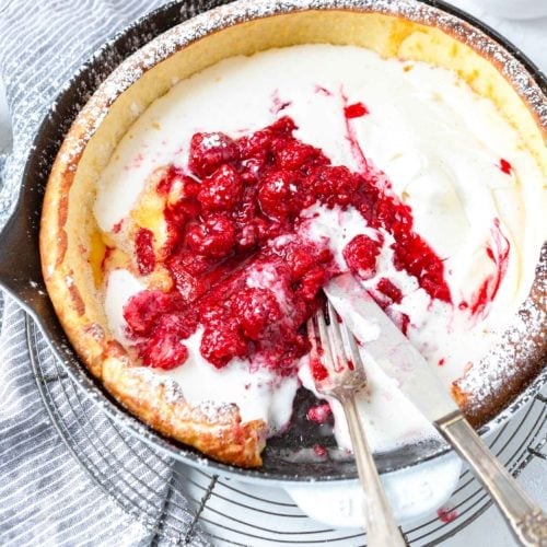 A photo of a dutch baby pancake in a cast iron skillet topped with whipped cream and fresh berries with one piece removed and a fork and knife laying on the side of the pan.