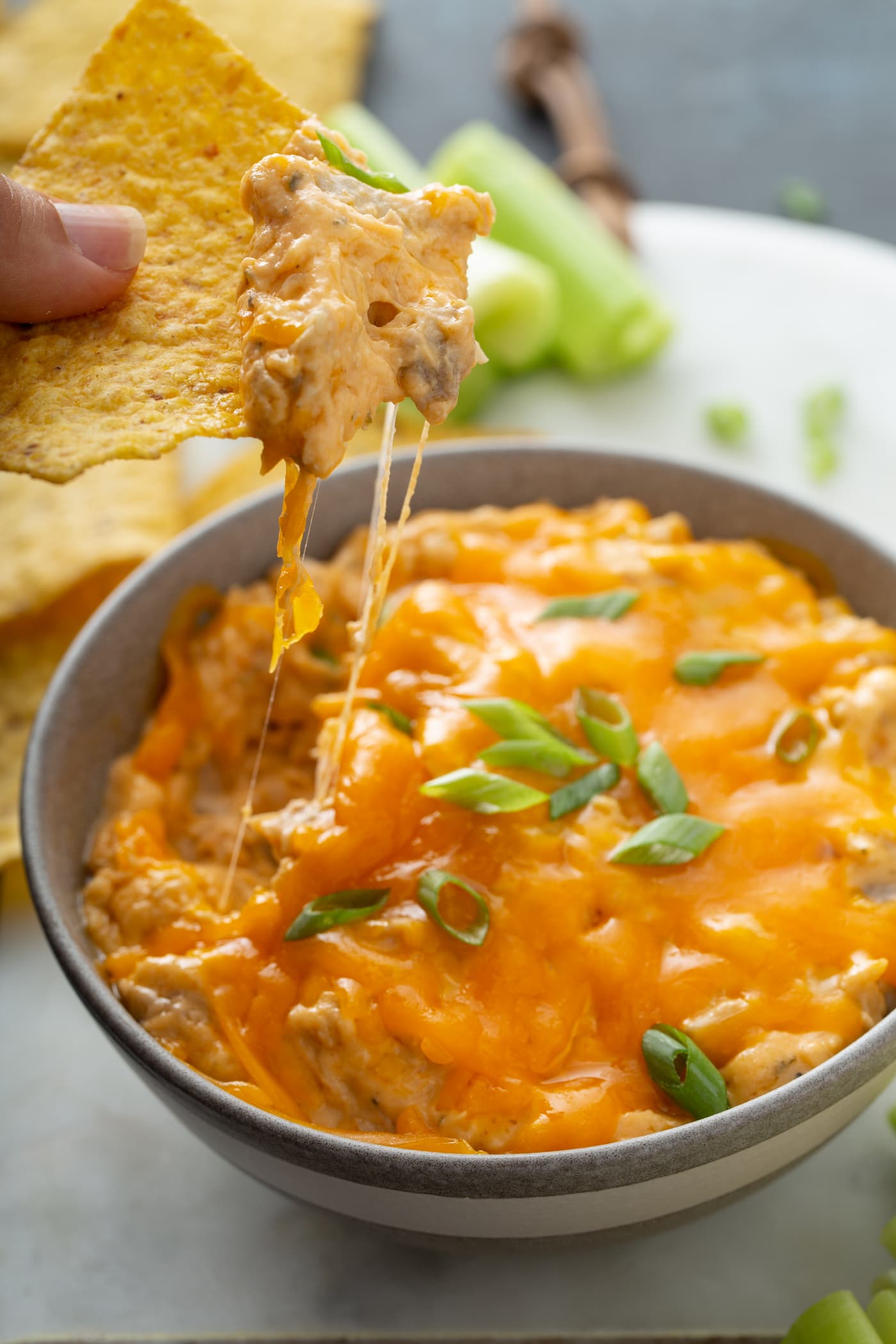 A bowl of buffalo chicken dip. A chip has been dipped in and melty stringy cheese is hanging from the tip of the chip. Chips and celery sticks are in the background.