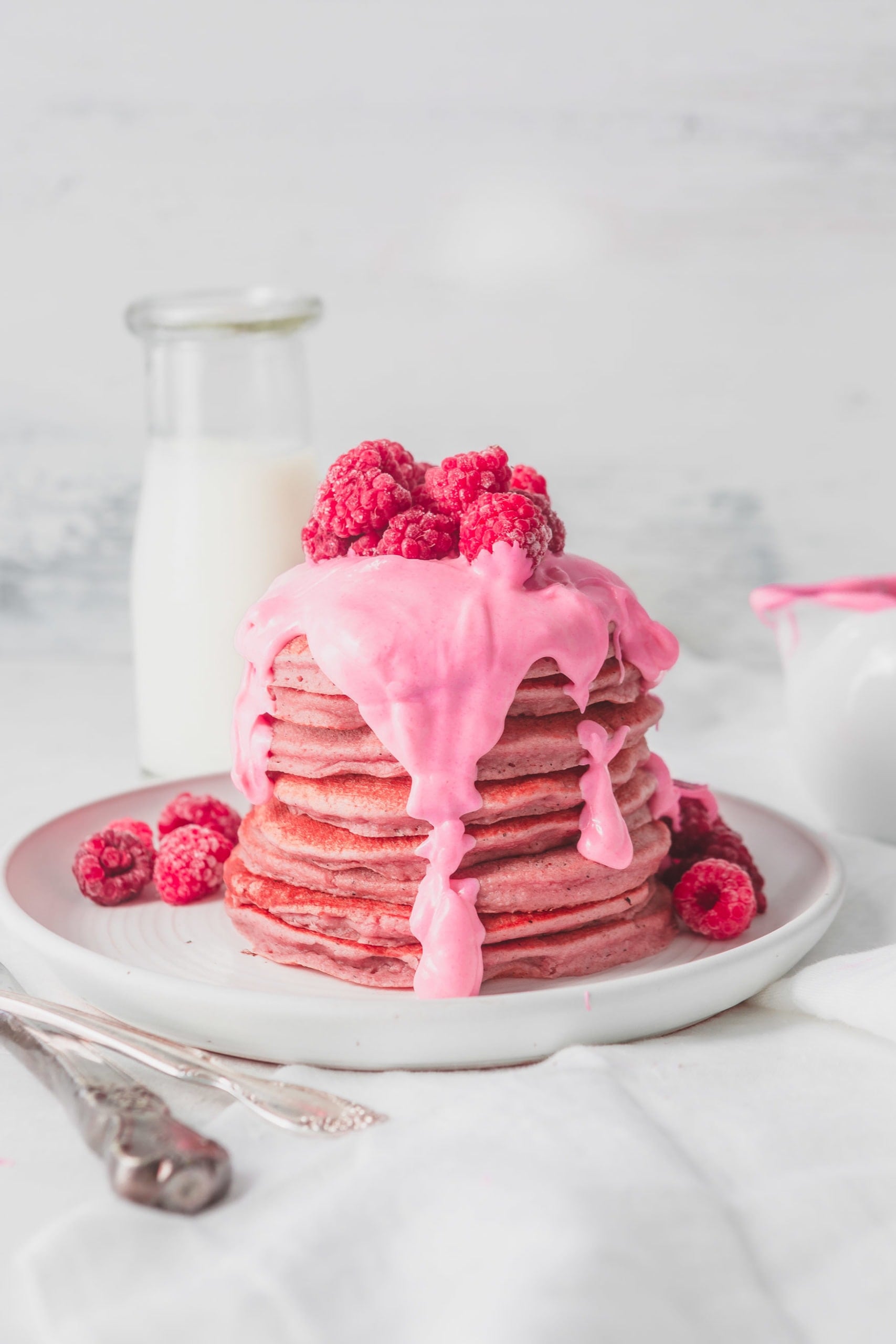 A large stack of pink pitaya pancakes with a thick pink chocolate sauce poured over the top. There are fresh raspberries on top of the sauce and raspberries on the plate around the pancakes.