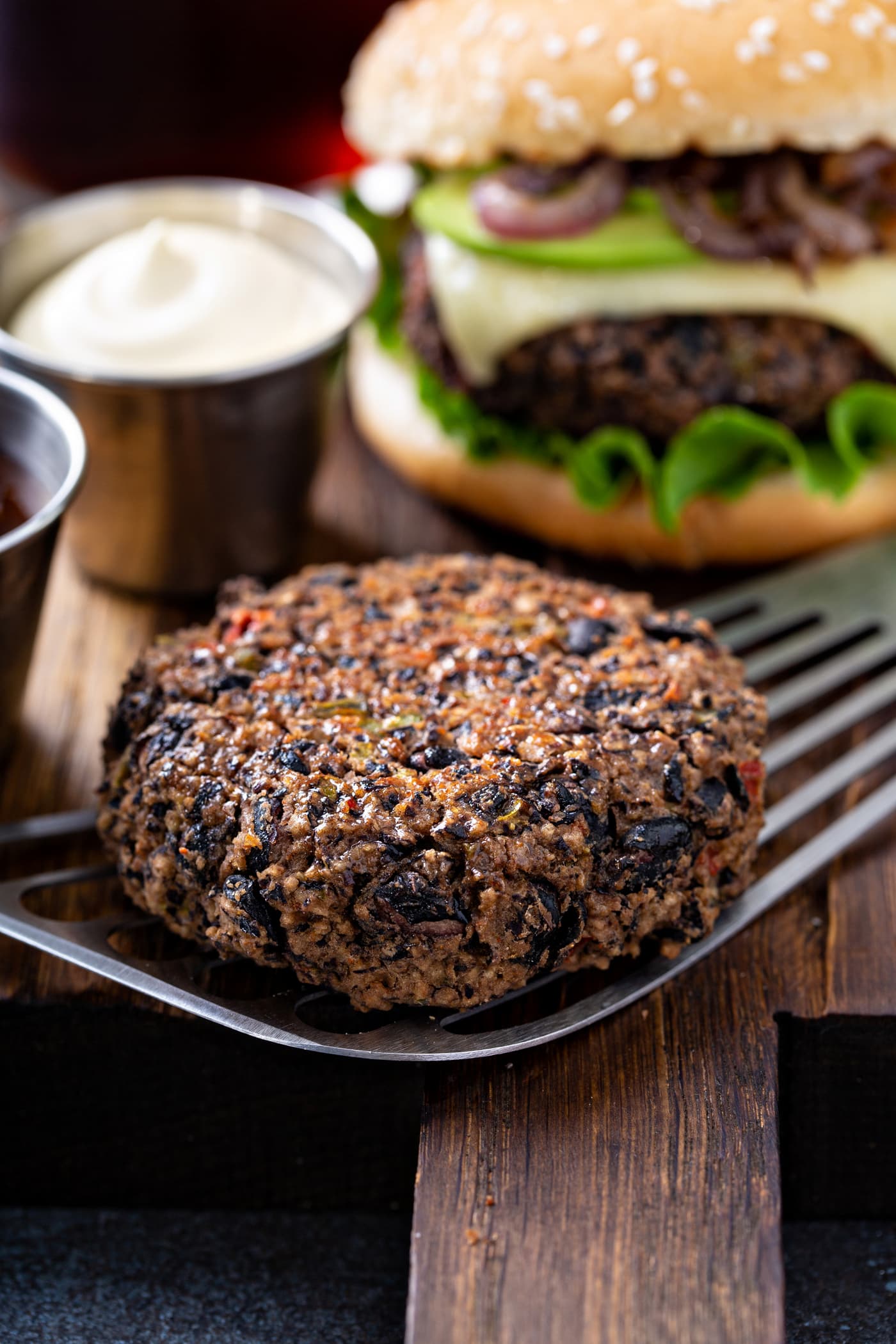 A cooked black bean burger patty on a spatula with a small cup of mayo in the background and a burger on a sesame seed bun dressed with lettuce, cheese, avocado and onions.