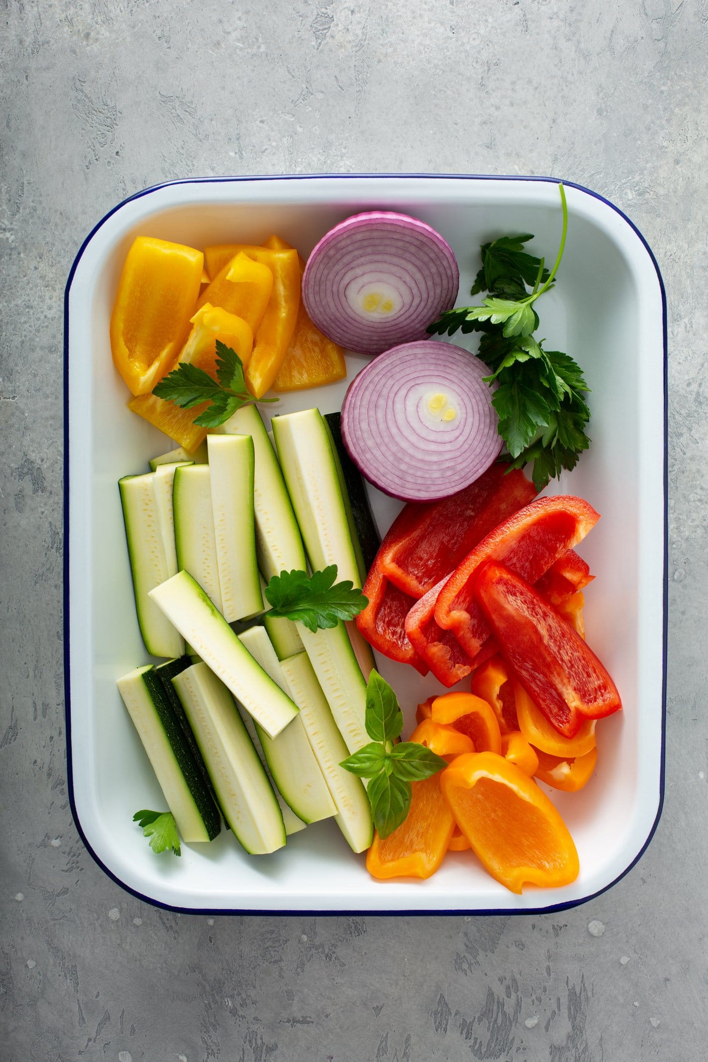 A plate with fresh zucchini wedges, sliced red onion, slices of red and yellow bell peppers, and parsley leaves.