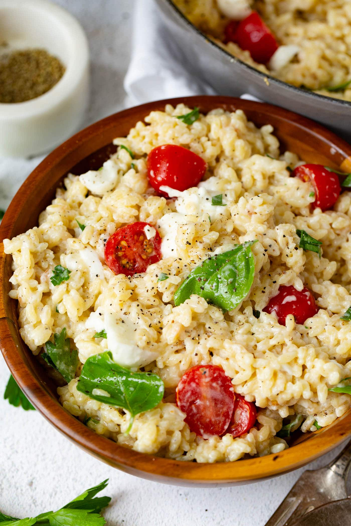 A bowl of caprese orzo salad. The salad has tomatoes, mozzarella chunks and fresh basil leave in it. A container of fresh ground pepper and a few parsley leaves are next to the salad.