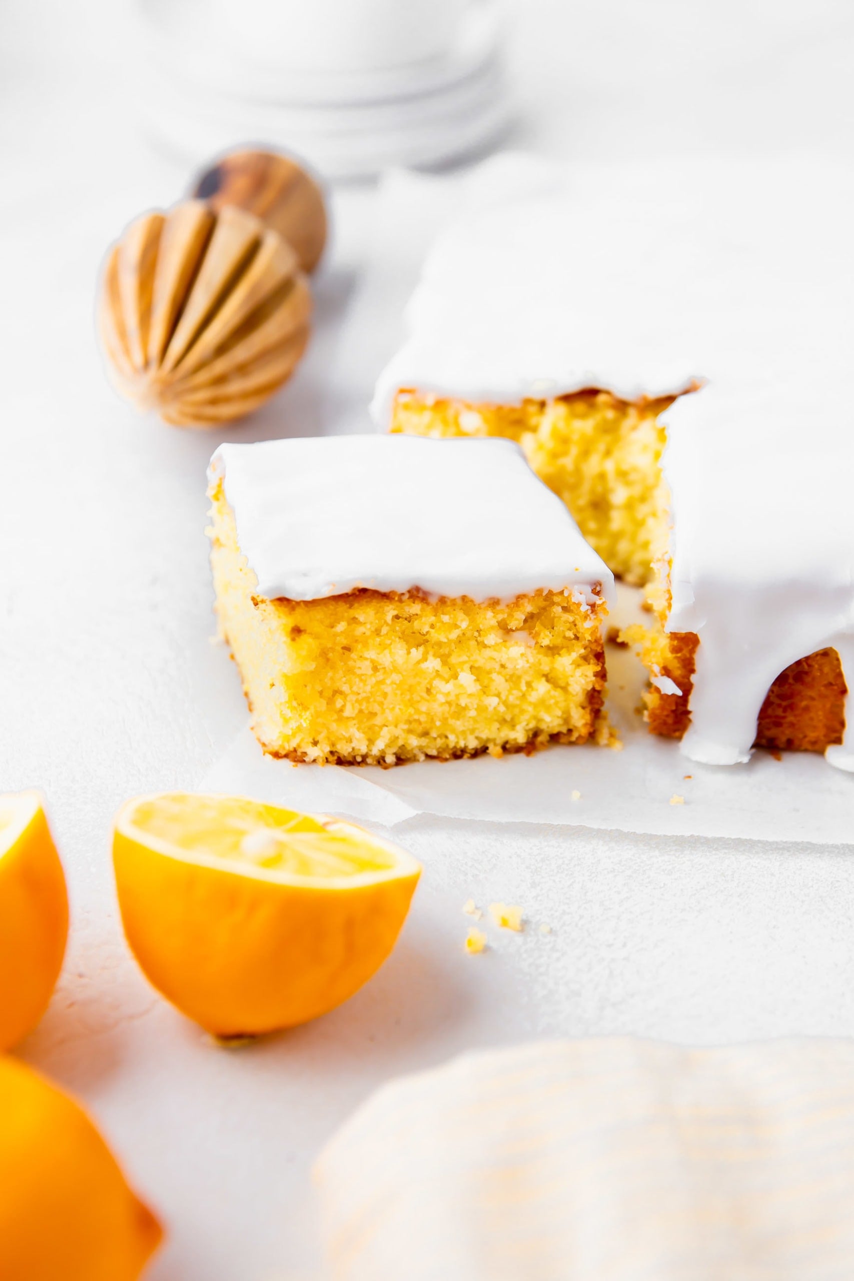 a photo of a piece of lemon cake that has been cut from a square lemon cake. it is covered in a thick white glaze. there is a half of a lemon in front of it.