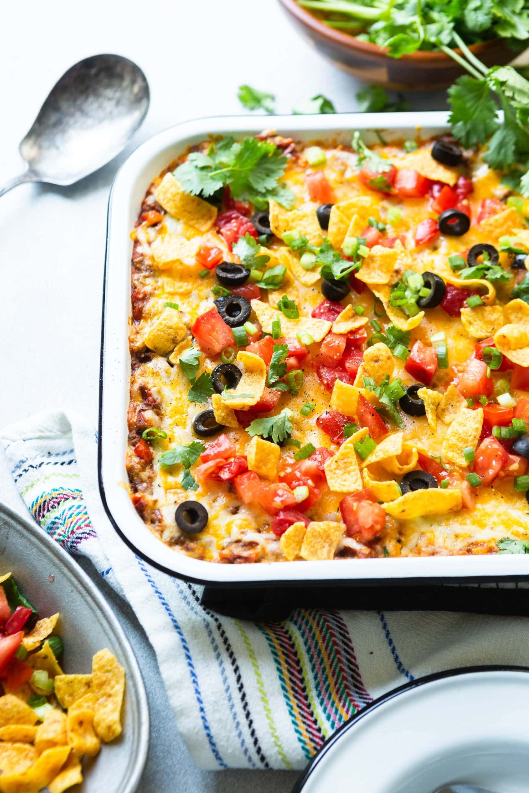 a photo of about half of a casserole dish full of taco casserole topped with fritos, black olives, diced fresh tomatoes, chopped cilantro.