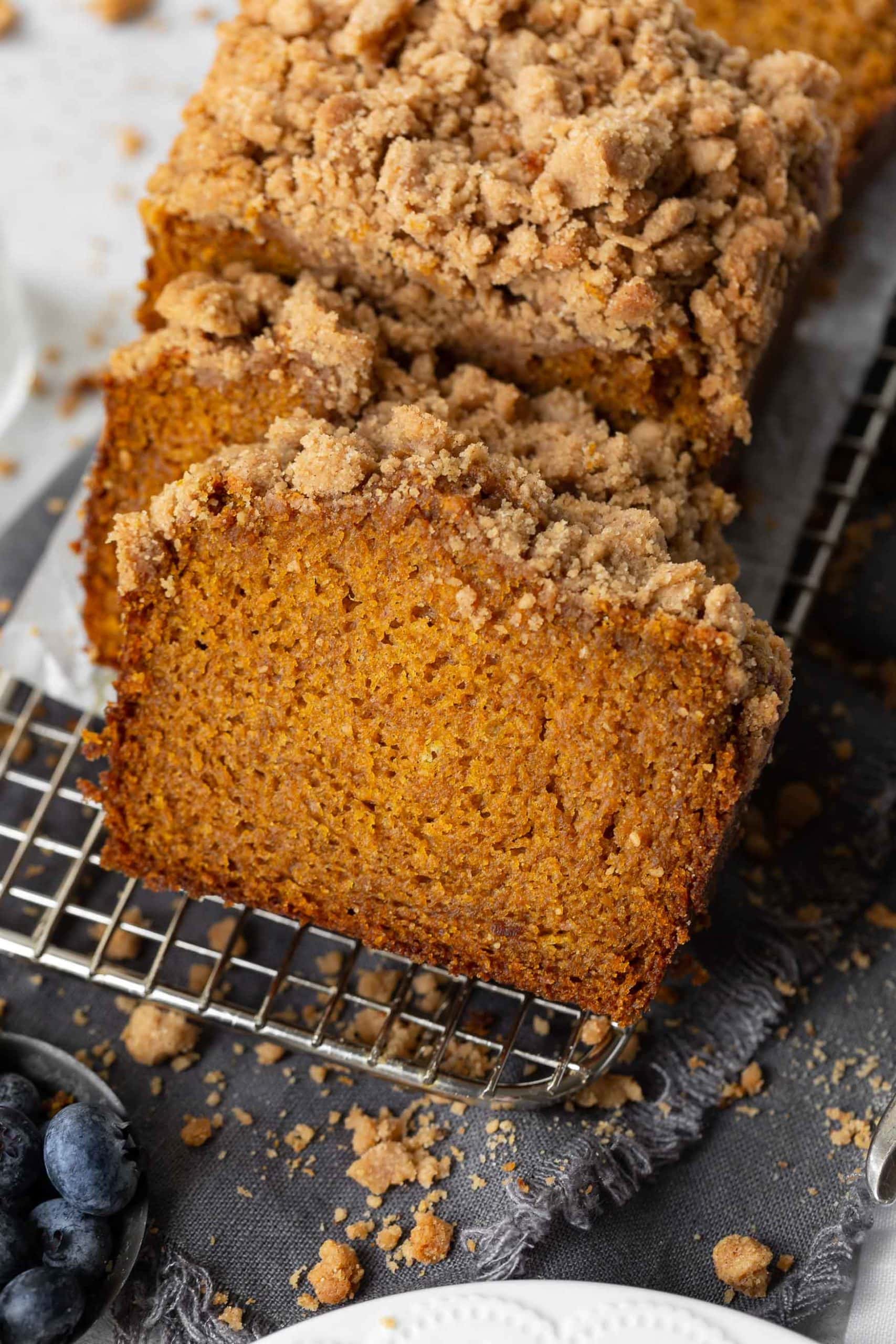 a photo of a loaf of pumpkin bread with cinnamon crumb streusel topping sitting on a narrow wire cooling rack. the loaf is sliced into individual slices.