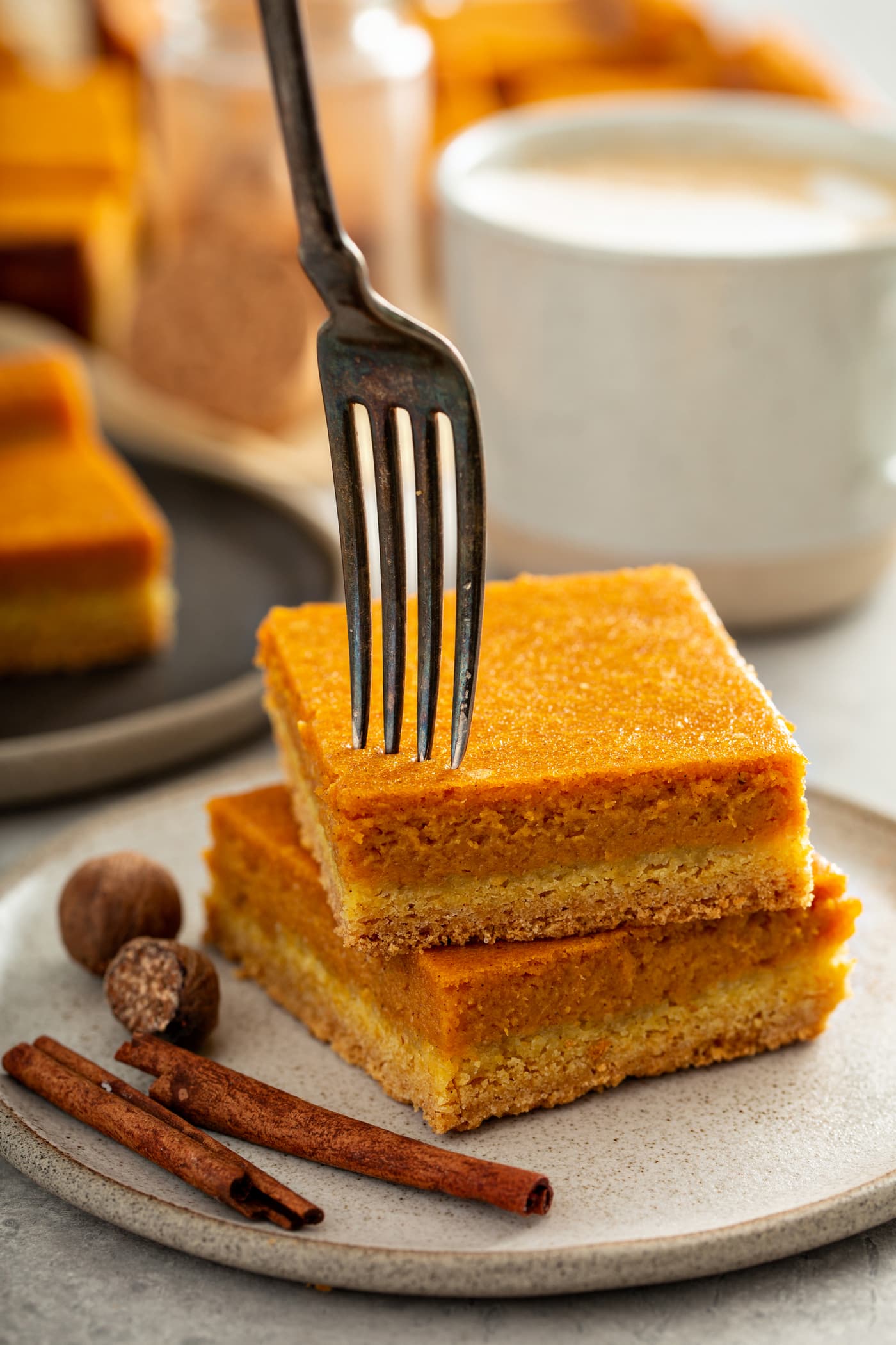 Two pieces of pumpkin cake stacked on top of each other. A fork is piercing the top of one piece. there are two cinnamon sticks and two whole nutmegs laying on the plate next to the cake. A plate of pumpkin cake and and white cup are in the background. 