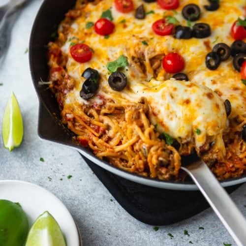 A cast iron skillet filled with cheesy ground beef taco spaghetti. A serving spoon is in the spaghetti and the dish is topped with melted cheese, tomatoes, olives, green onion and cilantro. Lime wedges are on the table next to the skillet.