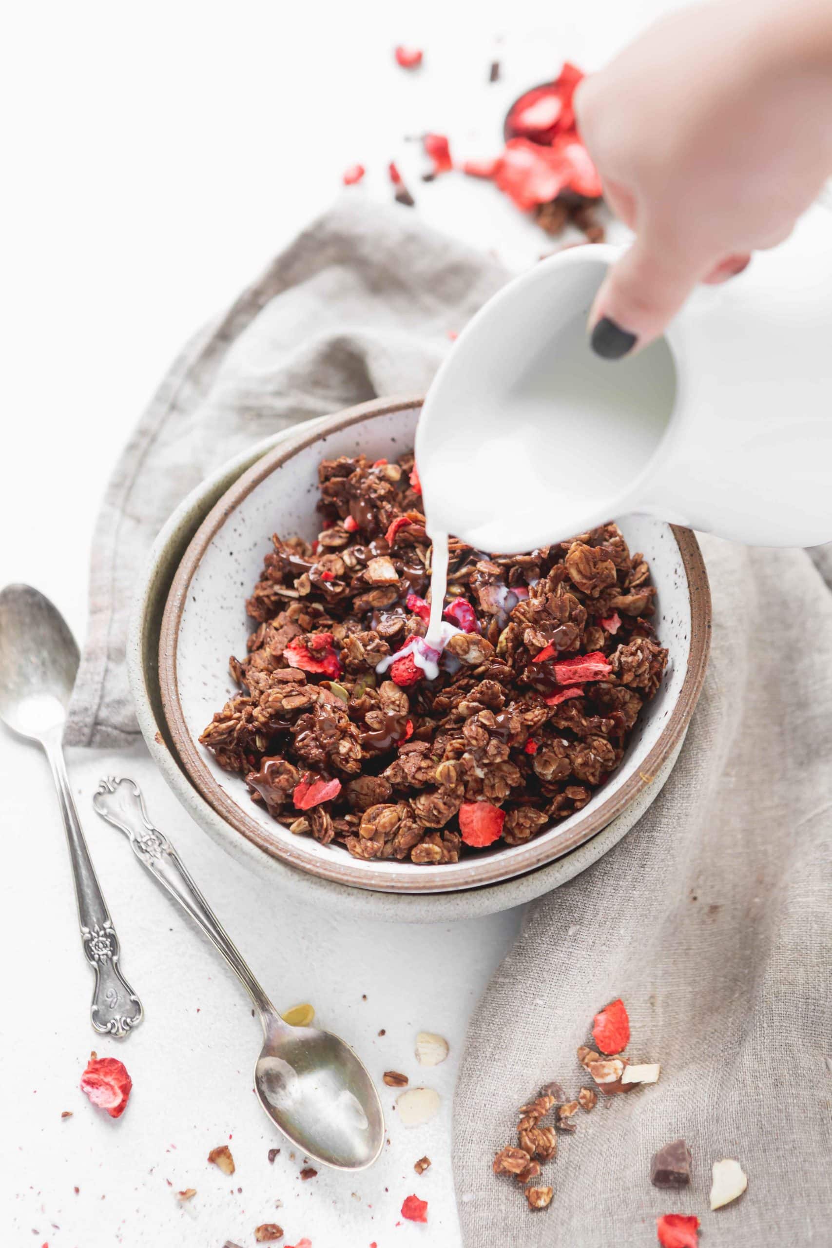A bowl of chocolate lovers granola with strawberries. Milk is being poured over the granola and two spoons are on the table next to the bowl of granola.