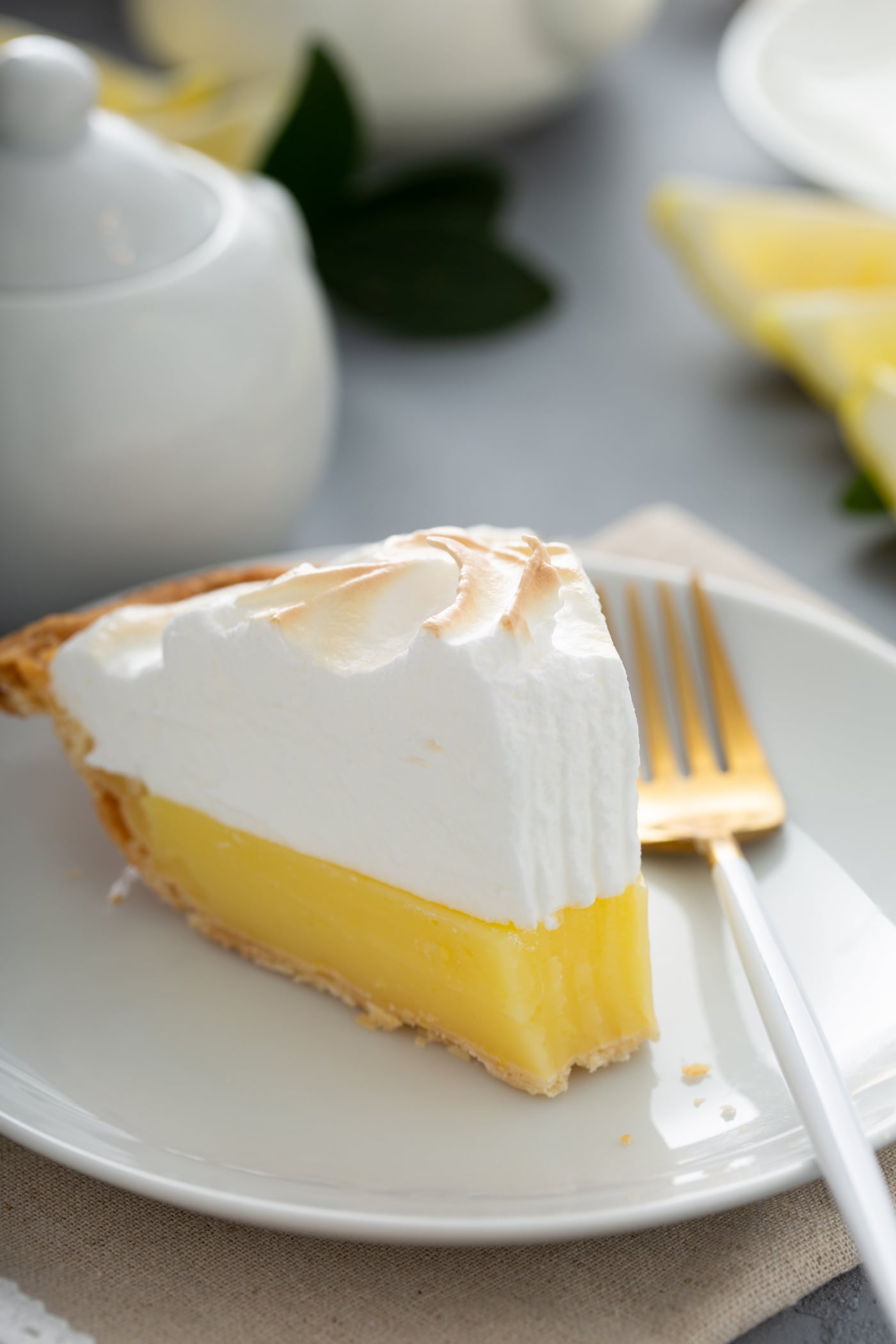 A slice of lemon meringue pie on a white dessert plate. A fork is next to the pie, and one bite has been taken from the slice.