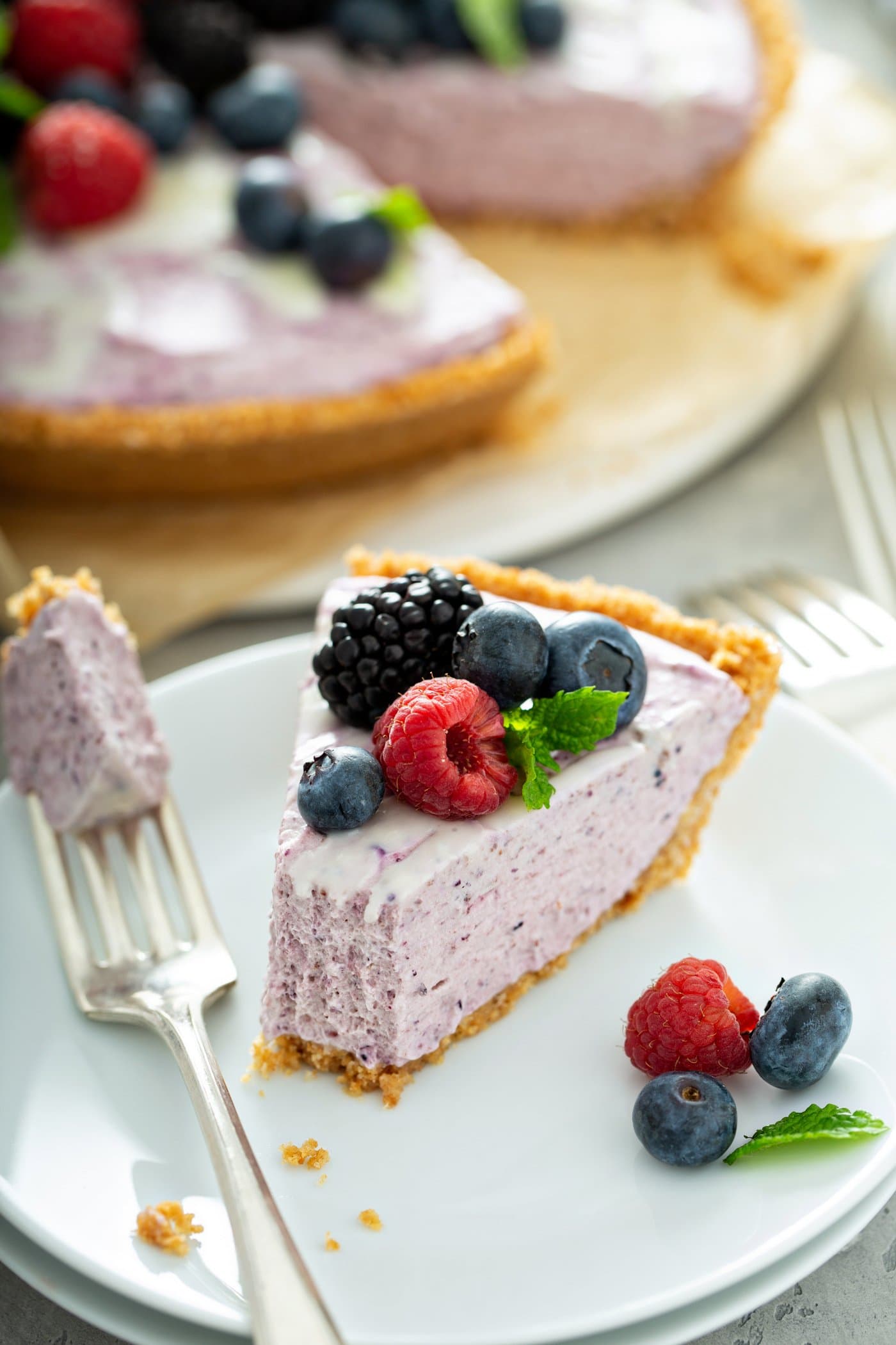A slice of white chocolate berry pie on a dessert plate. A fork with a bite-size piece of pie on it is next to the slice of pie. the pie is topped with raspberries, black berries and blue berries and one mint leaf. there are berries on the plate next to the pie and the whole pie in which a slice a has been removed, is in the background.