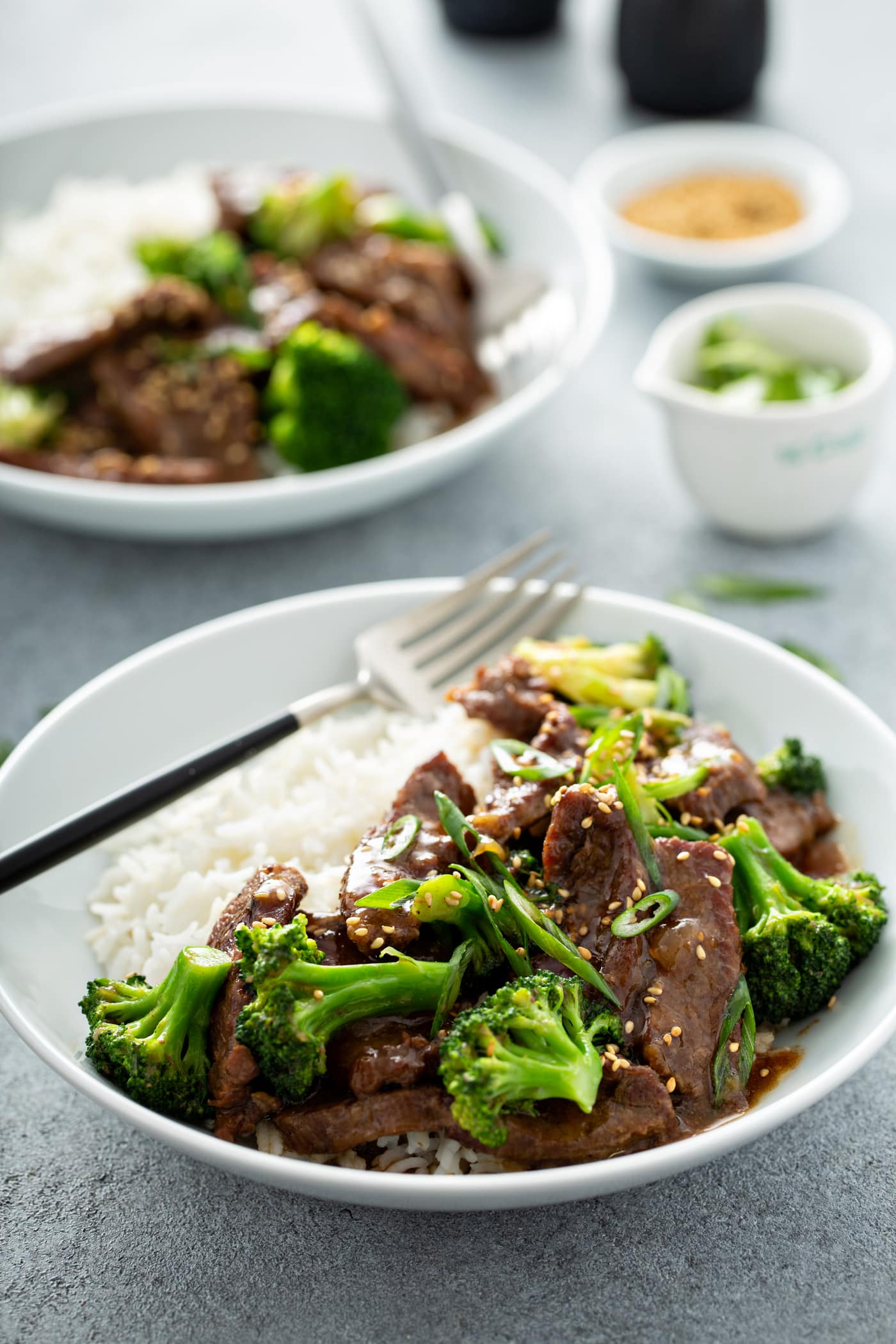 A photo of a bowl of beef and broccoli with another bowl of the same dish in the background.
