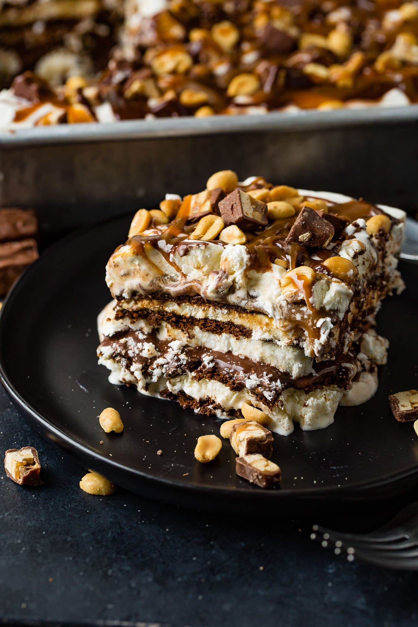A square piece of snickers ice cream cake on a brown dessert plate. There are layers of ice cream, chocolate cake, fudge, caramel and peanuts. There is fudge and whipped cream on top and salted peanuts are sprinkled over the whole thing. A pan of snickers ice cream cake is in the background.