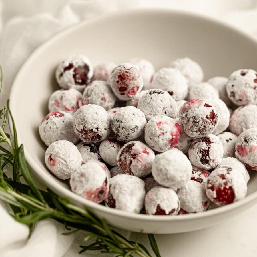 a small bowl of sugar coated whole cranberries with a sprig of fresh rosemary laying next to it. a small bowl of sugar coated whole cranberries with a sprig of fresh rosemary laying next to it.