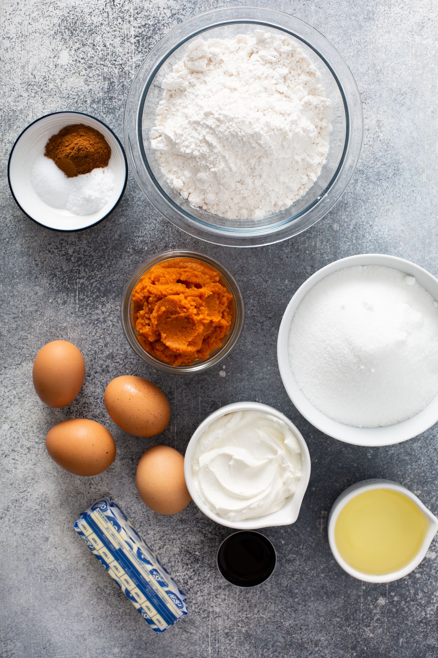 Small containers with ingredients for sour cream cinnamon swirl pumpkin bundt cake. There are containers of flour, sugar, sour cream, butter, pumpkin, and cinnamon. there are also four brown eggs on the table.