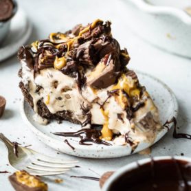 a photo of a slice of reeses ice cream pie sitting on a small plate topped with reeses peanut butter cups, and drizzled chocolate and peanut butter.