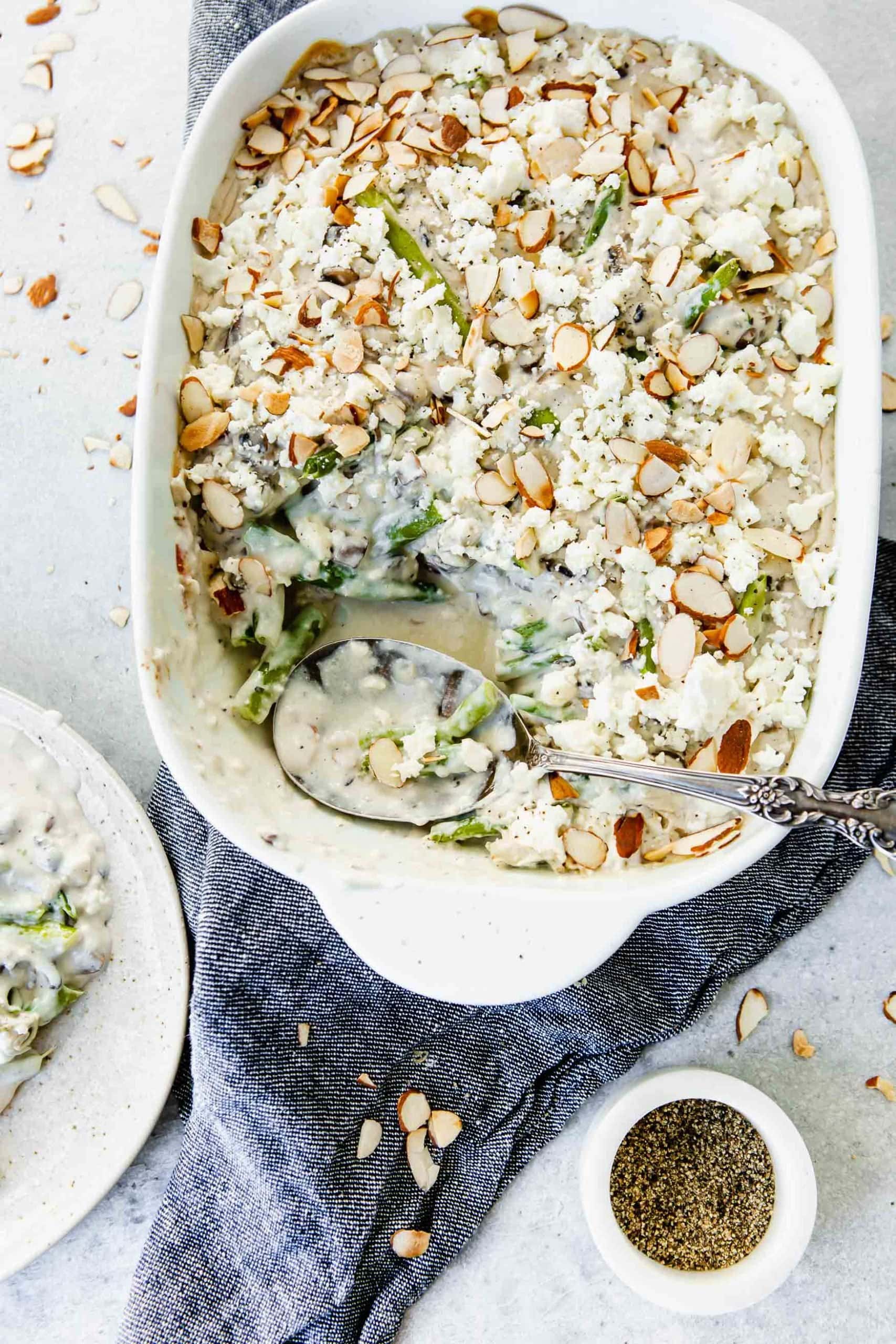 A casserole dish filled with green beans and feta. There is a serving spoon in the dish and you can see the creamy cheese sauce, green beans and sliced almonds on top. A dinner plate with a spoonful of beans is near the casserole dish.
