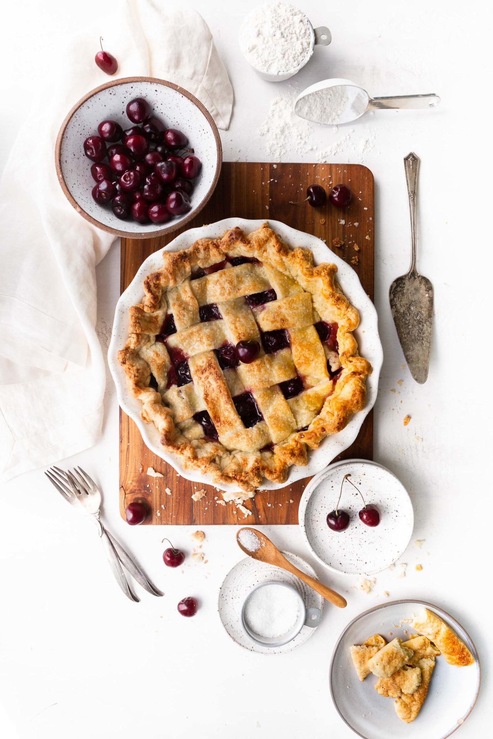 a photo of a whole cherry pie with a golden lattice top in a white pie dish sitting on a wooden cutting board surrounded by a bowl of fresh cherries, a silver pie server, a bowl of sprinkling sugar, and two silver forks.