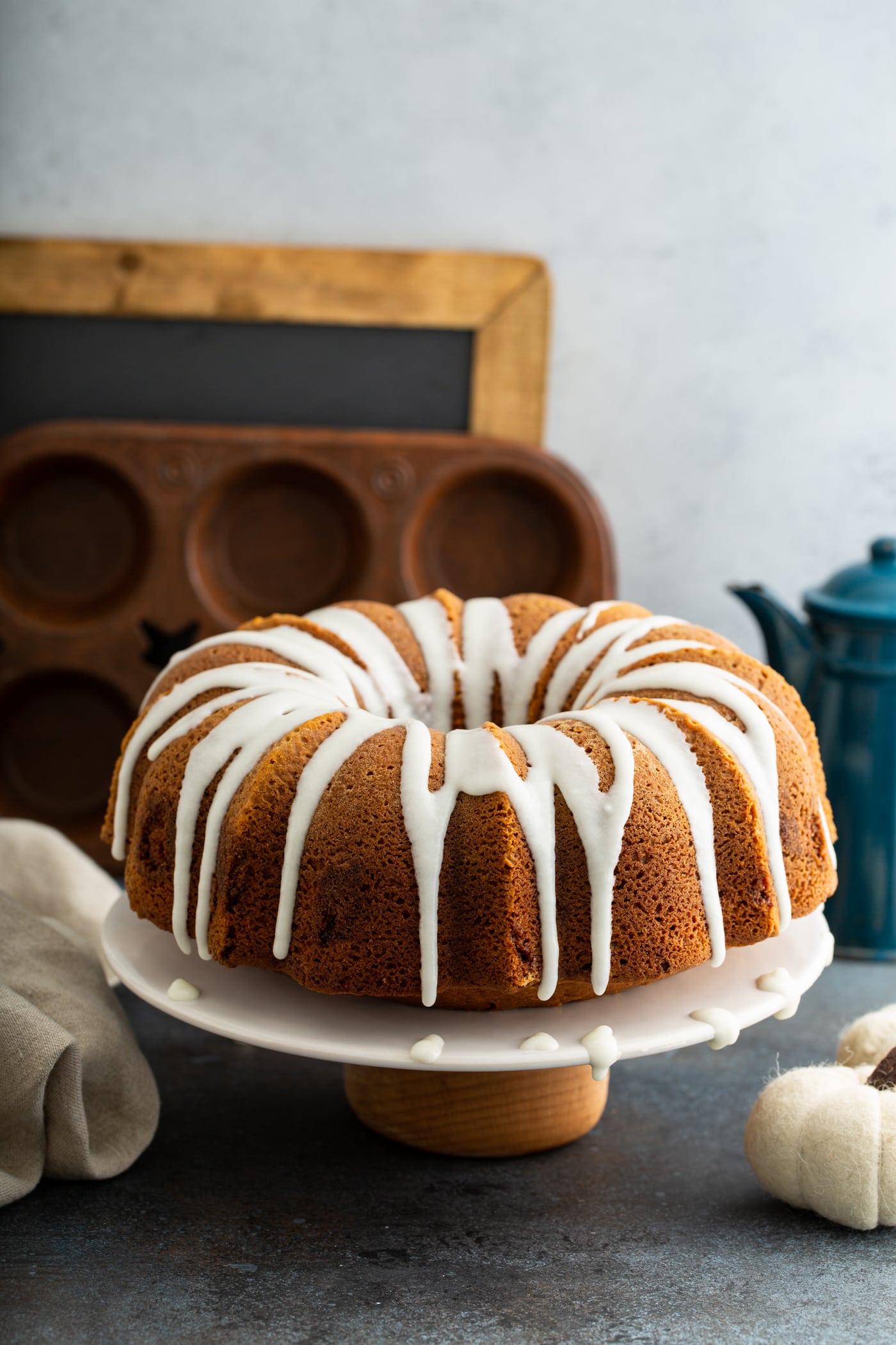 A sour cream cinnamon swirled pumpkin bundt cake with white glaze drizzled over the top. The cake is on a white cake plate and a muffin tin, chalk board, and coffee pot are in the background.