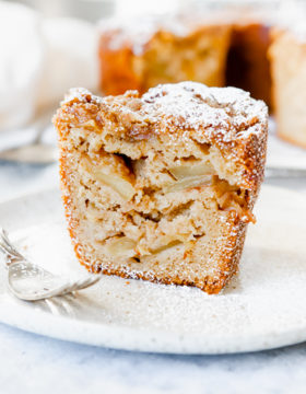 A slice of Jewish apple cake on a white plate with a fork. You can see the apples inside the slice. It has powdered sugar on top.