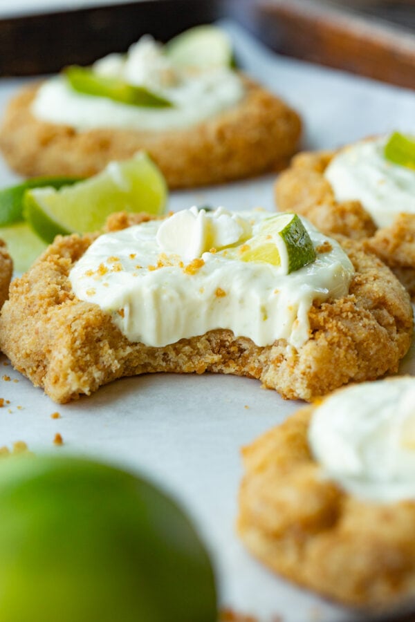 a photo of a key lime pie cookie that has had a bite taken out of it so you can see the creamy filling inside the graham cracker crust cookie topped with a small slice of fresh lime