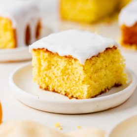 a photo of one piece of lemon cake topped with white frosting and sitting on a white plate. Several servings of cake are in the background.