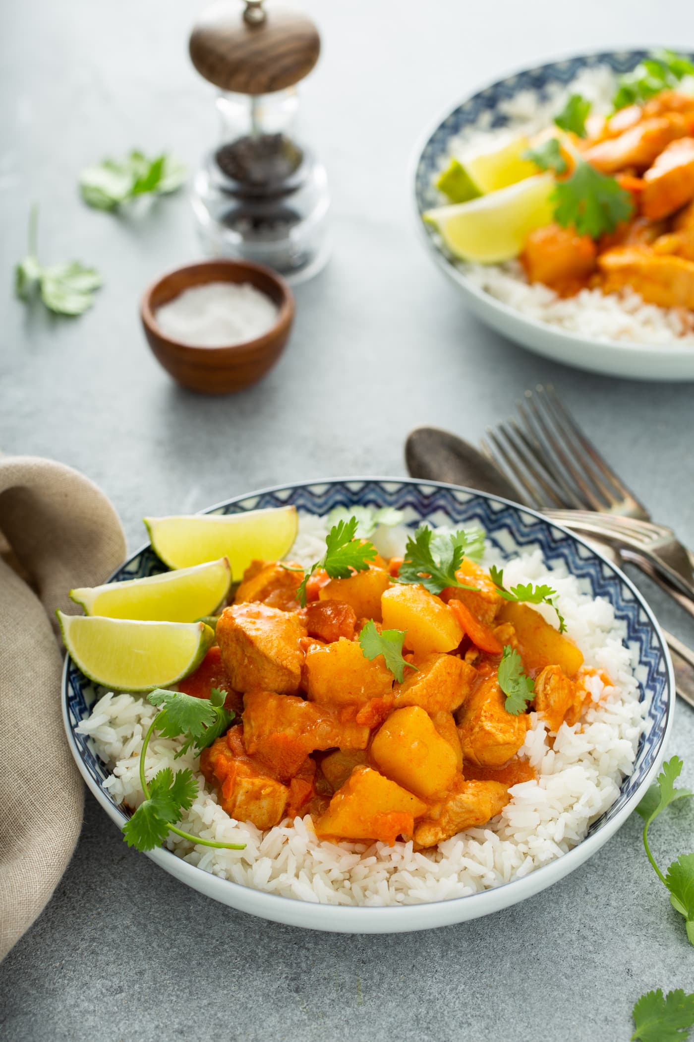 A bowl of white rice with chicken curry on top. There are chunks of chicken, potatoes and carrots in a curry sauce. Three lime wedges are on the edge of the bowl and fresh parsley is sprinkled on top. There are forks and spoons next to the bowl. In the background is a container of salt and pepper and another serving of chicken curry.