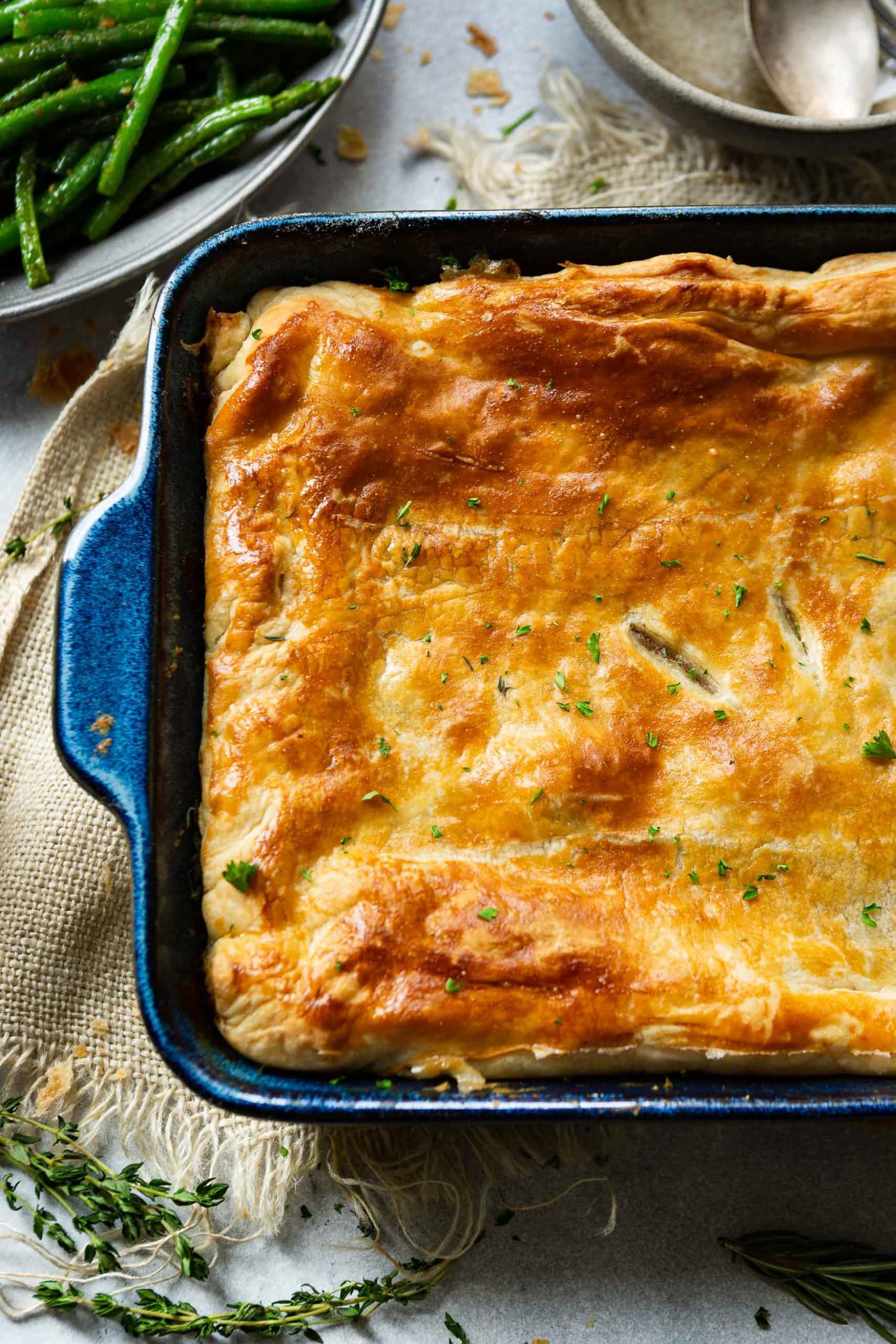 a photo of a dark blue rectangular baking dish will of beef pot pie topped with flaky golden puff pastry.