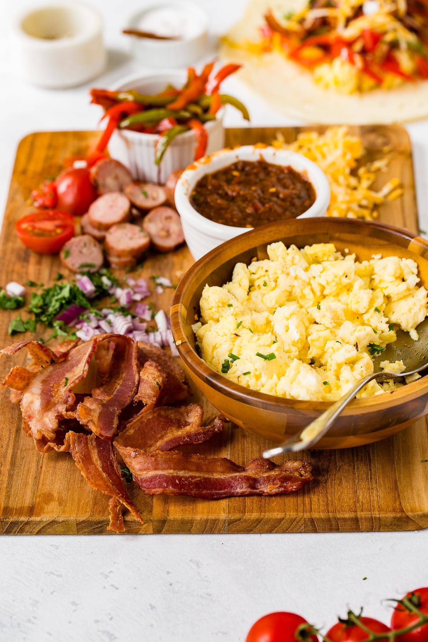 A wooden cutting board holding ingredients for apple chicken sausage burritos. There are sliced sausages, bacon, cheese, peppers, salsa, and a bowl of scrambled eggs with a spoon.