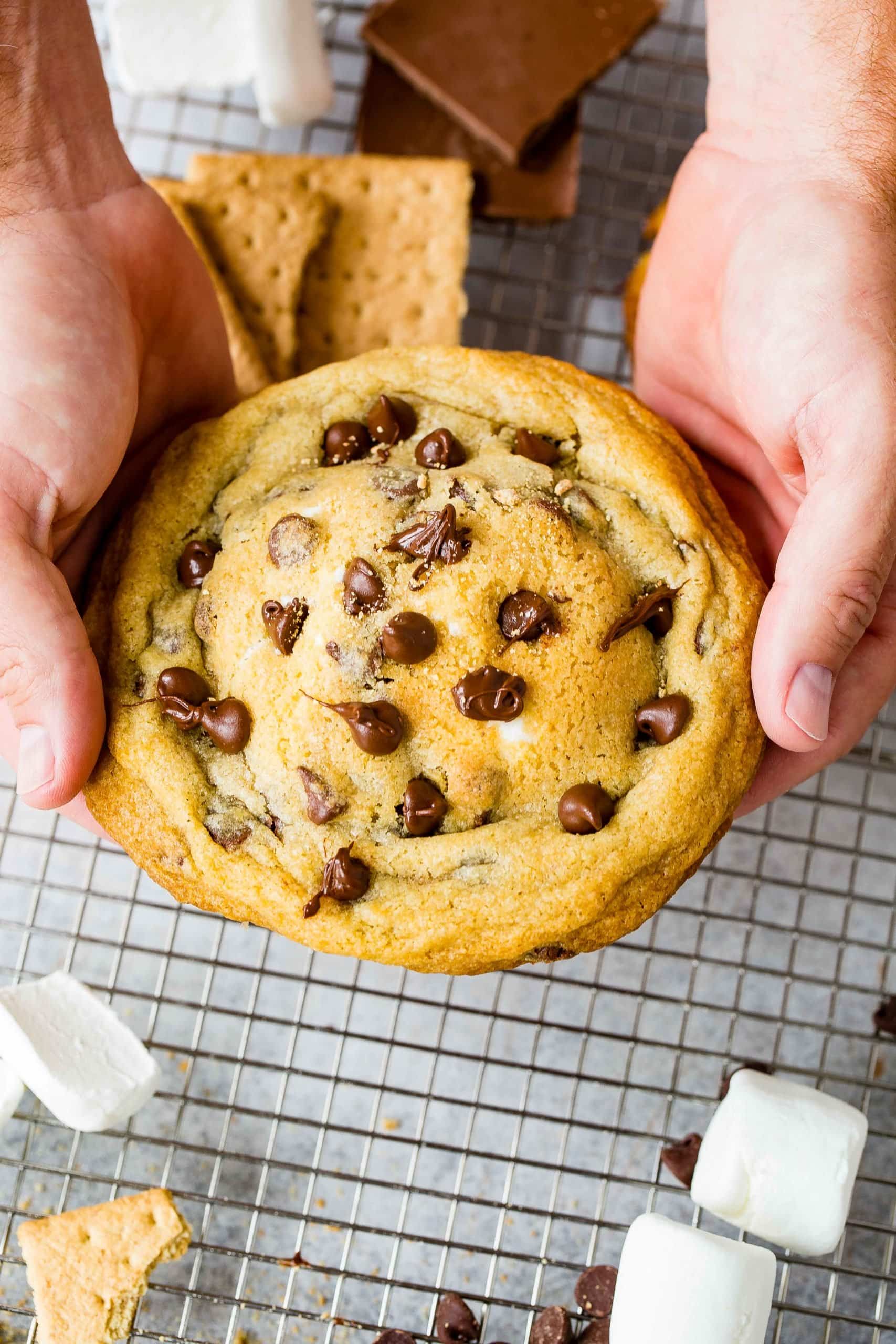 Two hands holding a large s'mores stuffed chocloate chip cookie. The cookie is loaded with chocolate chips and there are graham crackers and marshmallows on a cooling rack below the cookie.