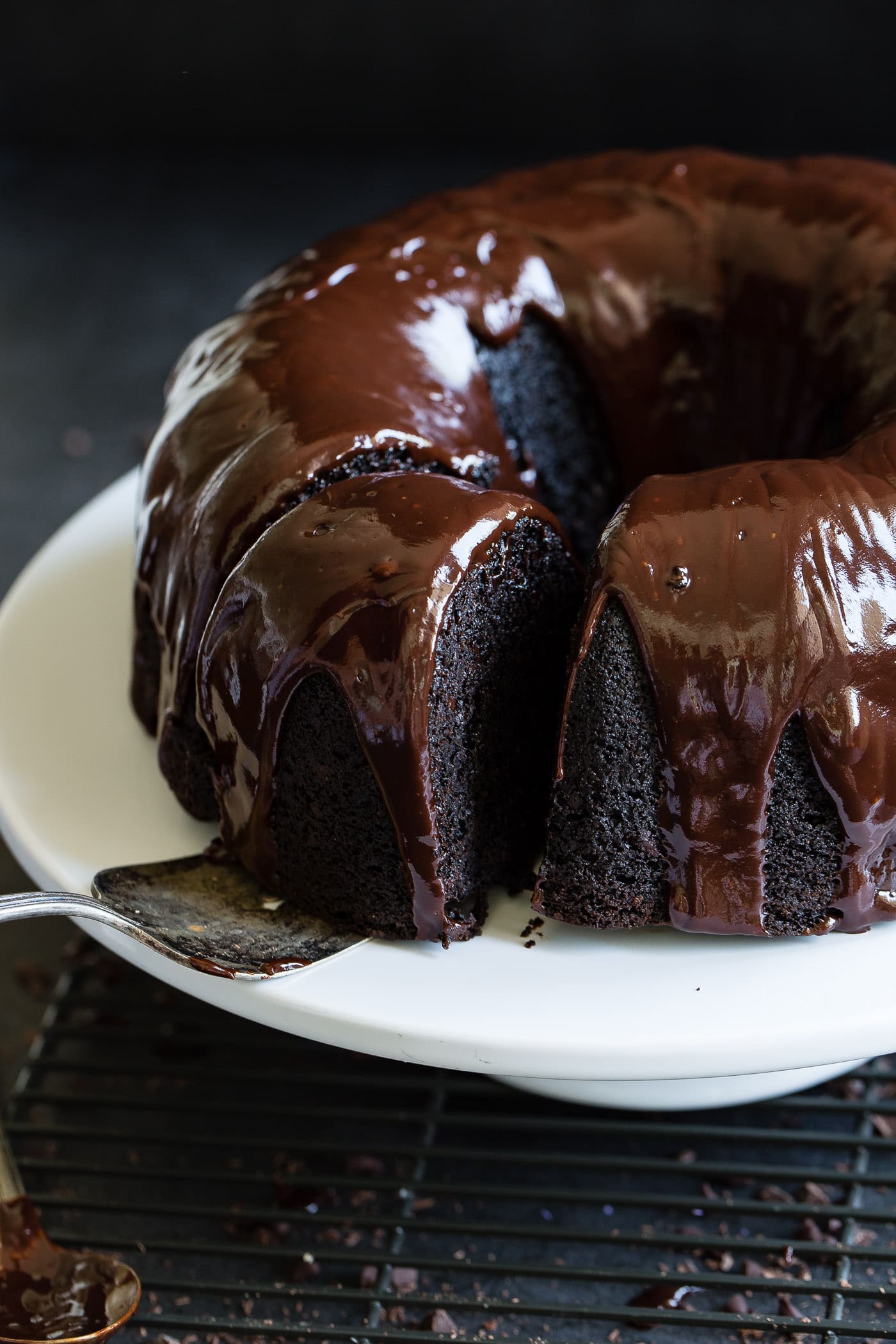 A slice of zucchini chocolate bundt cake being removed from the whole bundt cake. The cake has a chocolate glaze on it.