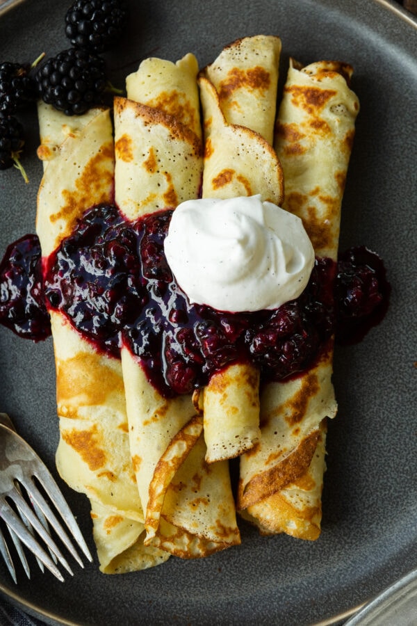 a photo of several golden pan fried thin finnish pancakes that are rolled up and topped with a blackberry sauce and whipped cream