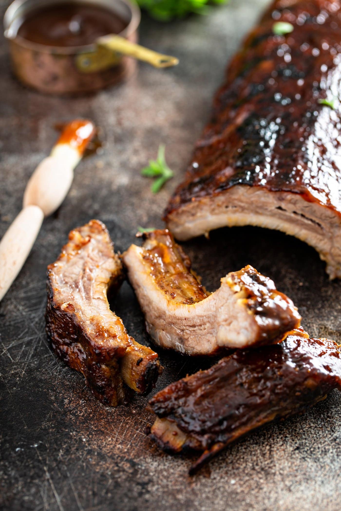 A rack of grilled pork ribs with a couple of ribs that are sliced off. A brush with glaze and a measuring cup of rub are in the background.