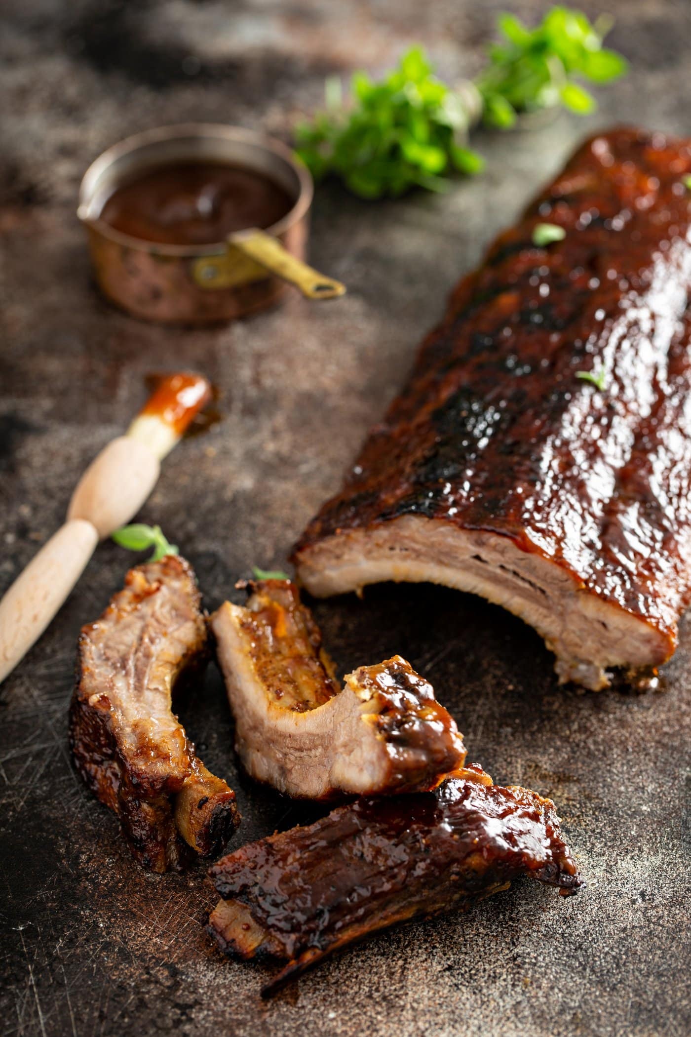 A rack of grilled pork ribs with a couple of ribs that are sliced off. A brush with glaze and a measuring cup of rub are in the background.