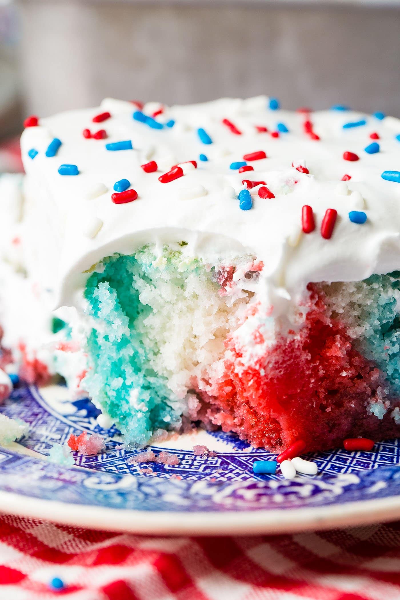 A piece of red, white, and blue poke cake. It is frosted with white frosting and sprinkled with red, white and blue sprinkles. The cake is white with areas of red and areas of blue.