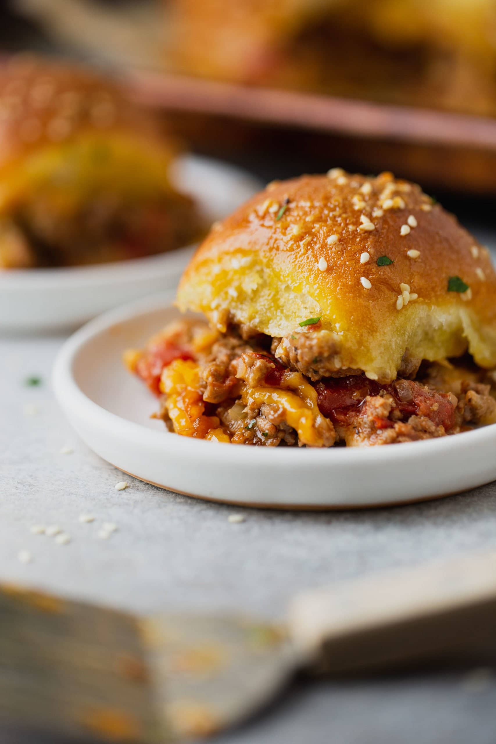 A cheeseburger slider on a plate. The golden roll is packed with ground beef, melted cheese and copped tomatoes.